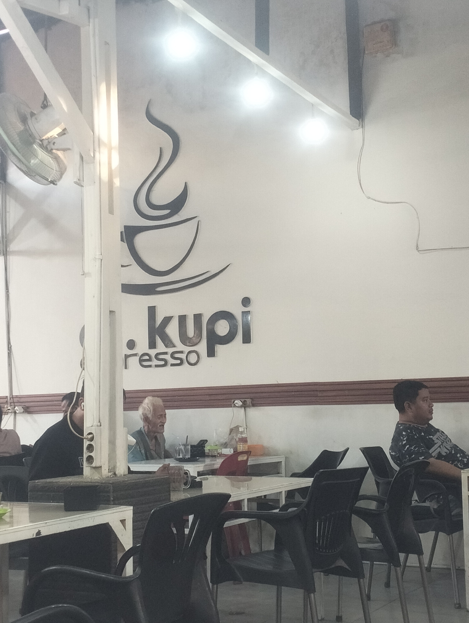 The image depicts the interior of a cafe in Lhokseumawe, Indonesia, identified by the logo ".kupi resso" on the wall. Three men are seated at tables. One man is wearing a black shirt, another man appears older with white hair, and the third man, on the right, wears a patterned shirt. The cafe has white walls with a dark brown trim, and overhead lighting. A ceiling fan is visible in the upper left corner. Tables and black plastic chairs are arranged in the foreground. A power outlet is visible on a vertical support structure.
