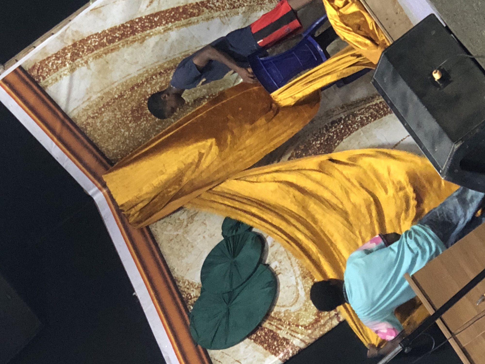 The image depicts an interior space in Onitsha, Nigeria, featuring two individuals, a yellow drape, a backdrop, a speaker, a green object and a blue plastic chair. One individual wears a blue shirt and red and black shorts, while the other wears a light blue shirt and jeans. The yellow drape is draped across the backdrop, which has a pattern of light and dark brown colors. A speaker sits to the side. A dark green object, composed of multiple lobes or layers, rests on the patterned backdrop. A blue plastic chair is positioned near the person wearing shorts.