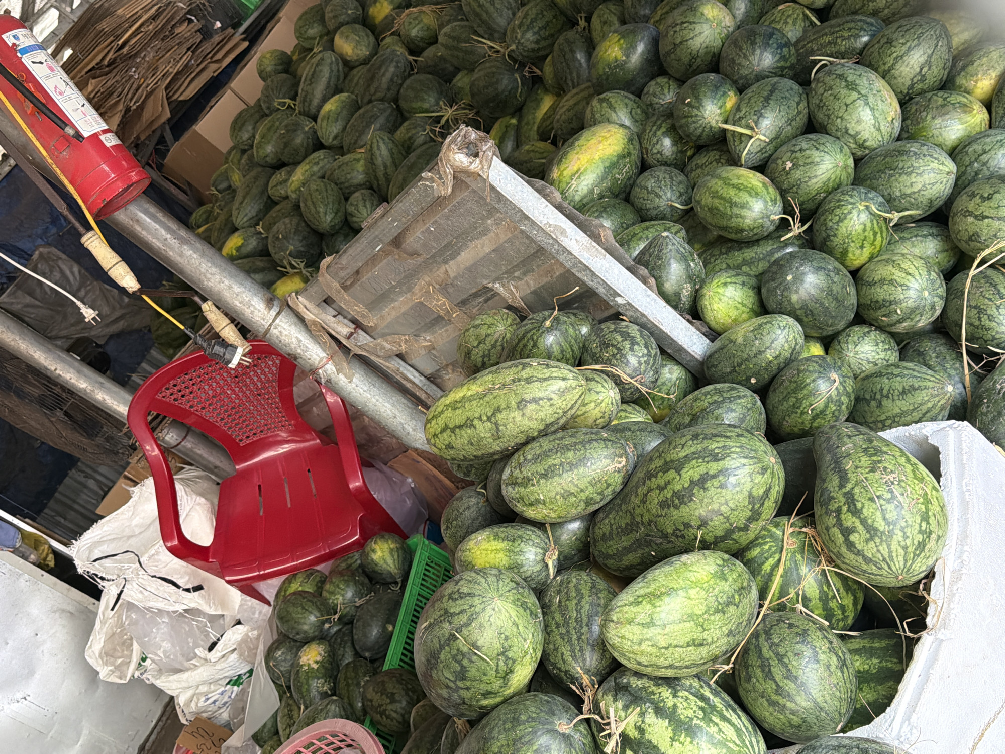 A street vendor is selling watermelons in Nha Trang Ward, Vietnam. The vendor has a large pile of watermelons, both large and small, arranged on the ground. There is a red plastic chair near the watermelons, and a fire extinguisher is mounted on a pole above the watermelons. A large white bag is also visible in the background. The scene appears to be set up in an open area, possibly a market or roadside stall.