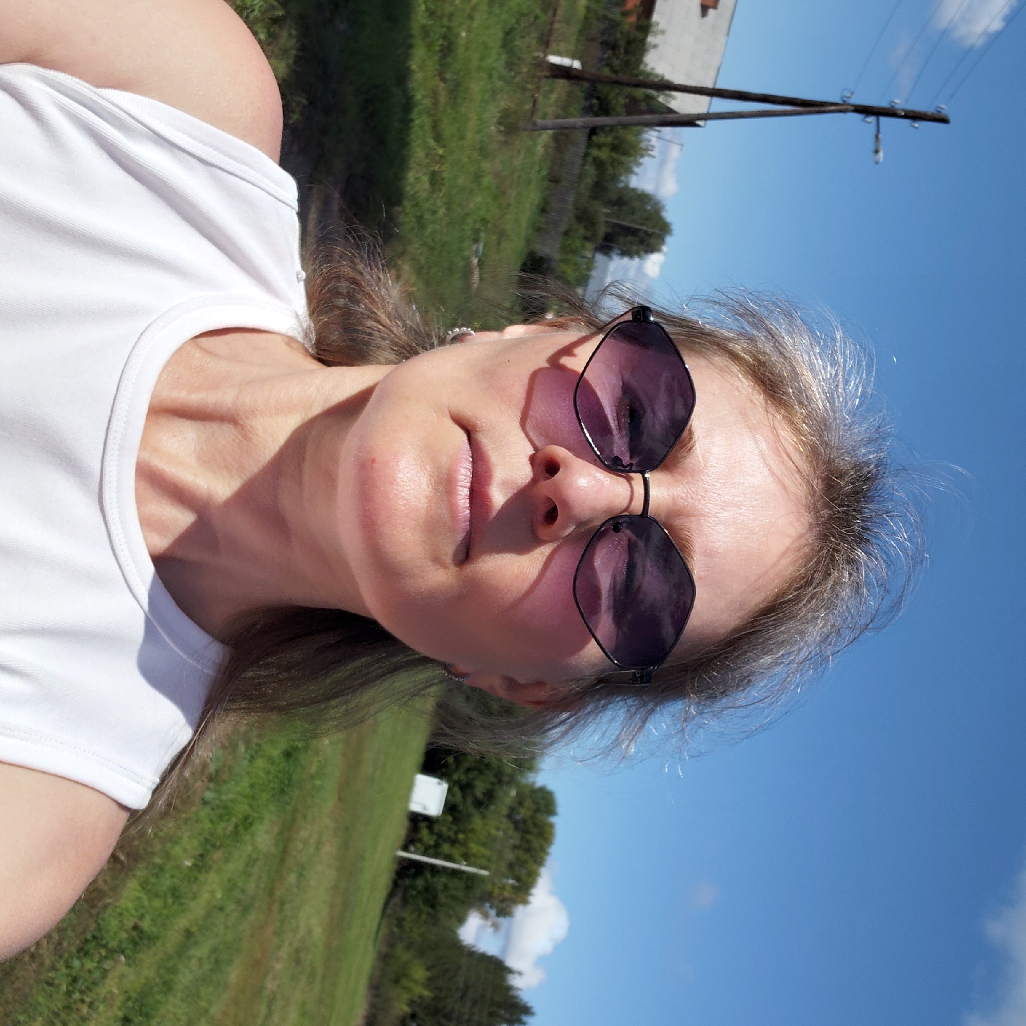 This is a selfie taken outdoors in what appears to be a rural or semi-rural setting, possibly the village of Novosyolovo, Russia, given the presence of a wooden utility pole and simple rural structures in the background. A woman with blonde hair, wearing sunglasses and a white tank top, is the primary subject. She is positioned in the foreground, her face is directed towards the camera. Behind her, a landscape of green grass, trees, and a bright blue sky with scattered clouds is visible. A wooden utility pole stands to the left, and some buildings and a fence are discernible in the middle ground. The lighting suggests it is daytime, with shadows cast on the woman's face and neck.