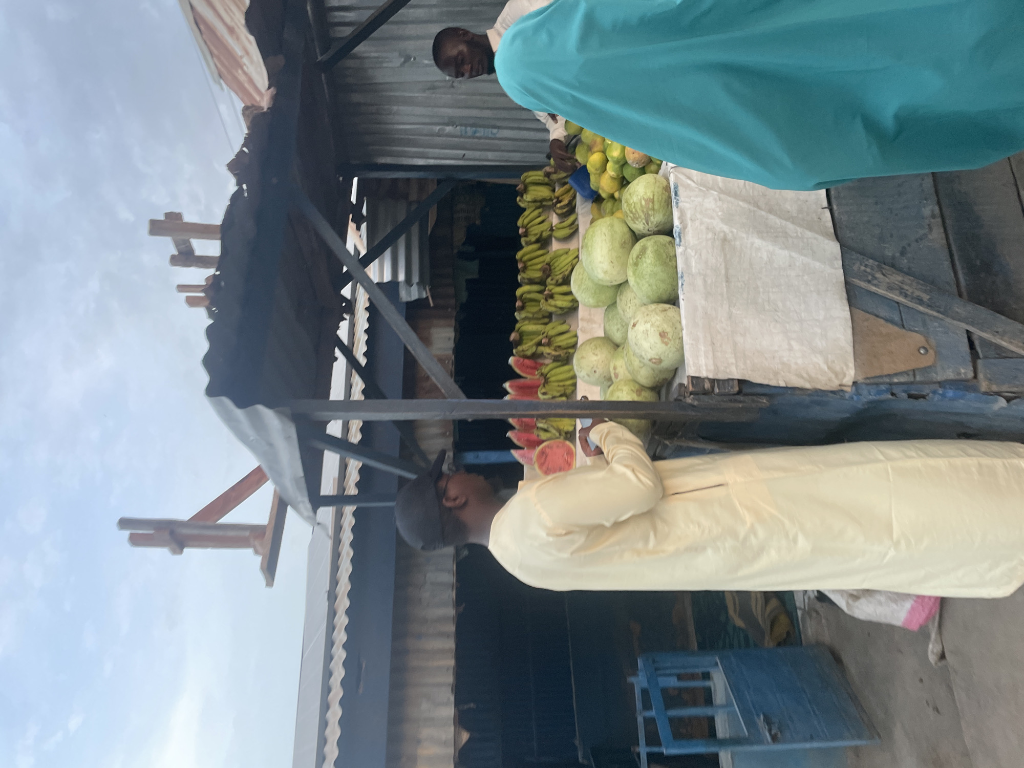 A fruit stand in an open-air market. Melons, bananas, and mangoes are displayed. A man in a light yellow robe is examining the watermelons. Another man is standing behind the fruit stand, wearing a blue garment. The structure of the market stall is visible, made of metal and wood. The sky is visible in the background.