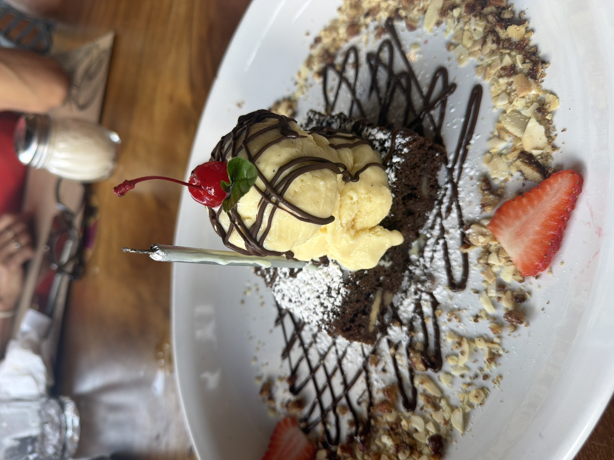 A top-down, close-up shot shows a dessert dish on a white plate. The main components are a brownie, topped with a scoop of vanilla ice cream, drizzled with chocolate syrup, and garnished with a maraschino cherry and a small green leaf. Powdered sugar is sprinkled on the brownie. Sliced almonds and chopped nuts surround the dessert. A sliced strawberry is also present. A silver utensil lies next to the cherry. In the periphery, a hand and part of a salt shaker are visible, suggesting a dining context. The scene's location is likely Puebla, Mexico.