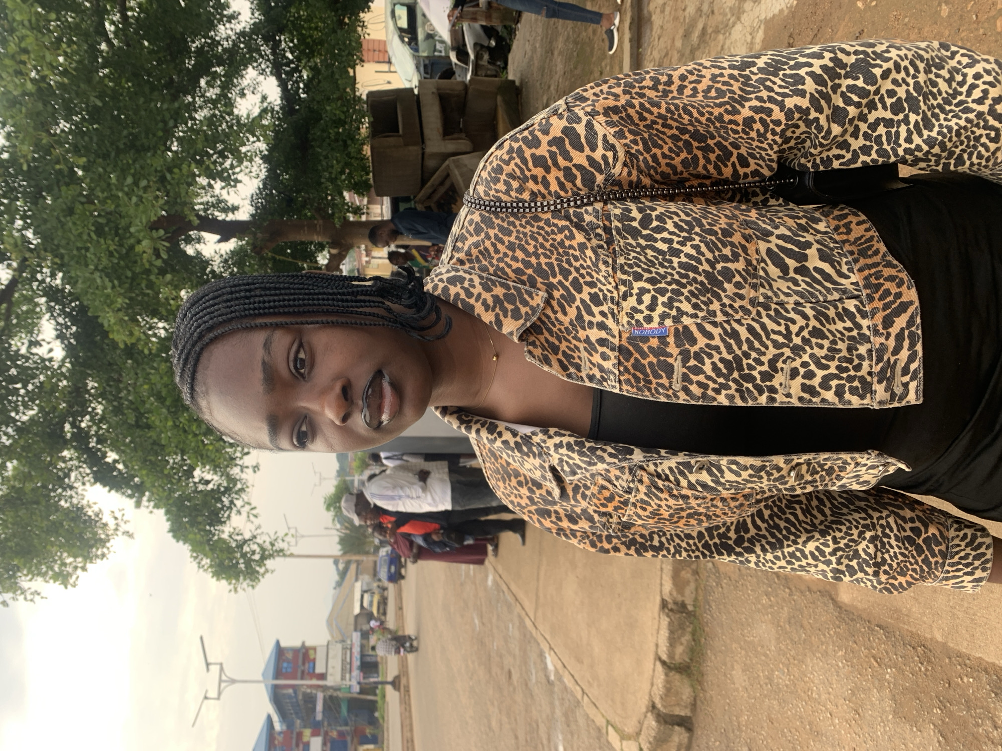 A young Black woman with braided hair and dark lipstick stands on a sidewalk, looking to her left. She wears a leopard-print denim jacket over a black top and a black skirt. In the background, several people stand near a road, with vehicles and buildings visible. A large tree with green leaves is behind the woman. The environment appears to be a street scene in Rafin Par, Nigeria, with the characteristic cloudy sky and a dusty road. There are some concrete blocks and possibly abandoned car parts to the right of the woman.