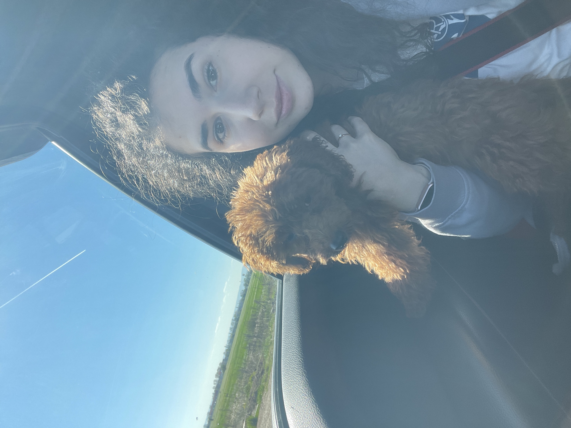 A woman with long curly brown hair is in a car with a brown poodle, smiling at the camera. The dog is partially hanging out of the car window. The car is driving past a green field under a clear blue sky with a visible contrail. The setting appears to be rural and there are no indications to suggest this is in Sofia, Bulgaria.