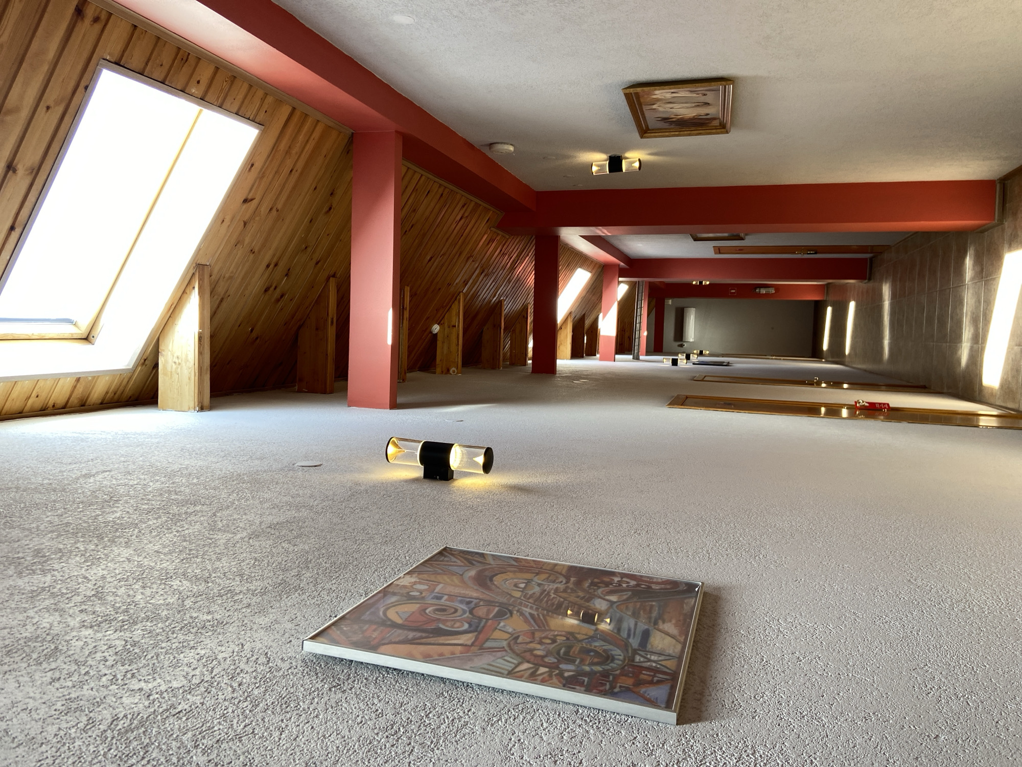 This media file captures a large, empty indoor space, likely a renovated attic, loft, or former commercial hall, in Kaliningrad, Russia. The room is characterized by its mixed architectural elements and abundant natural light.
On the left, a sloped wall is clad in light-toned wooden paneling, featuring several large skylight windows that allow bright daylight to illuminate the area. The ceiling is white, contrasted by prominent red structural beams and pillars that define the space and extend into the mid-ground. The floor is covered with a light grey, textured carpet or similar flooring.
In the foreground, a framed abstract artwork lies flat on the floor. Near it, a modern wall sconce, detached from a wall and placed on the floor, emits a warm yellow glow. Further back, a traditional framed painting is mounted on the ceiling. The distance reveals more red beams and pillars, and a tiled wall on the right with additional light sources casting bright strips onto its surface. A white air conditioning unit is visible on a far wall.
The absence of people and the arrangement of objects suggest the space is in a state of transition—perhaps undergoing renovation, being prepared for an event, or recently vacated. The strong natural light indicates it is daytime. There is no visible text.