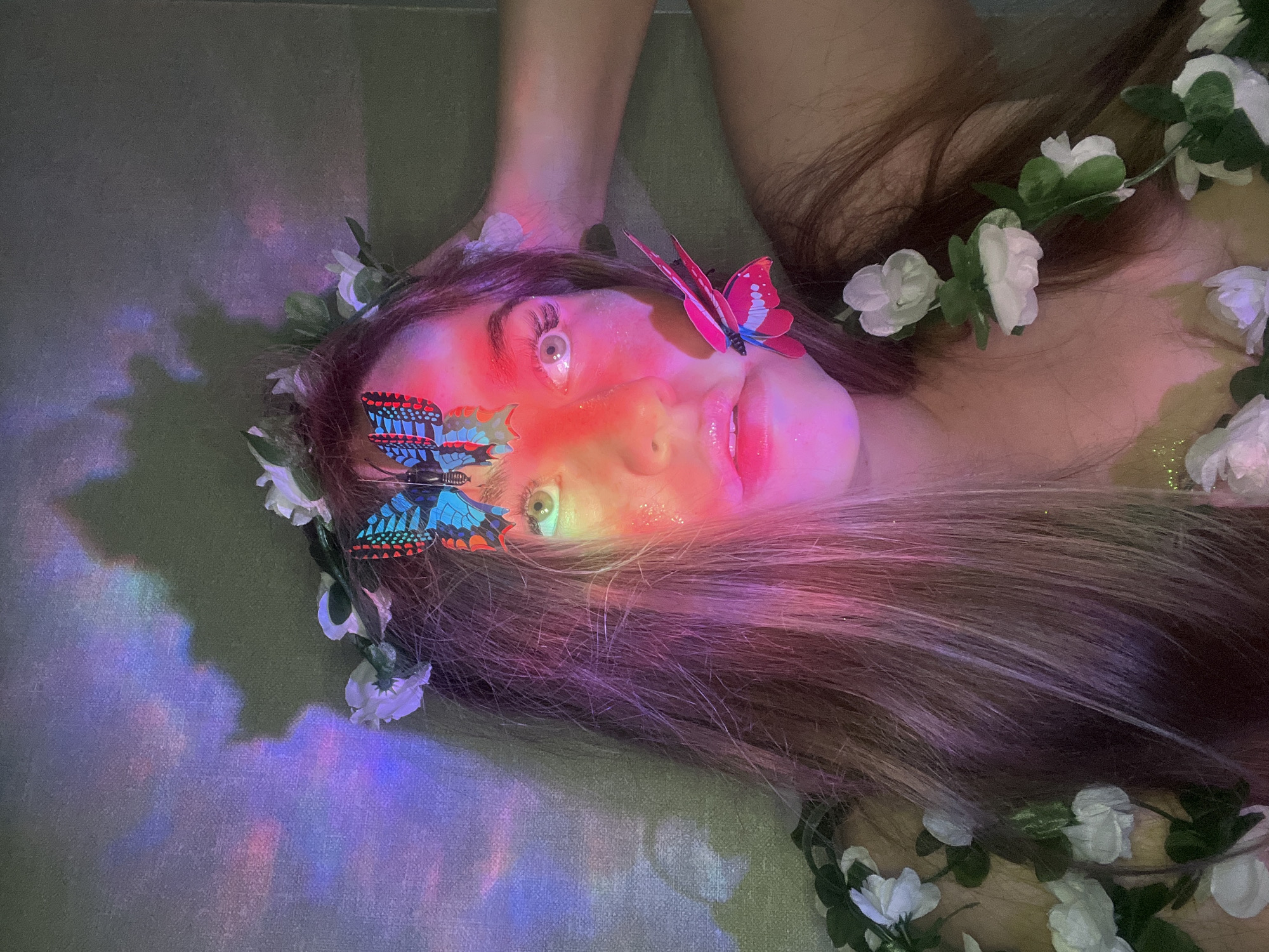 A woman is lying down, facing upwards. She has long brown hair adorned with a flower crown of white flowers and green leaves. There are two artificial butterflies attached to her face, one blue and one pink. Her face is illuminated with a rainbow-colored light. The location appears to be indoors, potentially in Chaikovsky, Russia. There are no visible actions or interactions depicted.