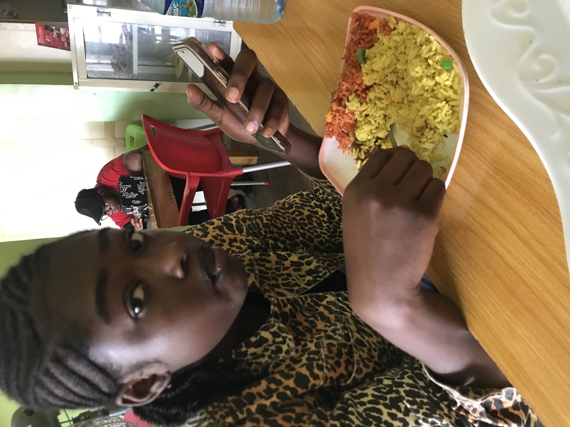 An indoor scene within an eating establishment in Kabong, Nigeria, features two individuals. In the foreground, a young Black individual with dark skin and braided cornrows styled diagonally is seated at a light brown wooden table, looking directly at the camera. They wear a leopard print jacket over a dark shirt. Their right hand holds a silver smartphone with a gold/bronze case, while their left hand holds a fork, poised over a rectangular pinkish plate of food. The plate contains two distinct sections of rice: yellow rice mixed with green peas and small orange pieces, and reddish-orange rice with similar orange pieces. A plastic bottle of Owan Spring Water with a blue label is positioned on the table to the right of the plate.
In the background, a second Black individual, wearing a black baseball cap and a red short-sleeved t-shirt, is seated at another wooden table, looking down and interacting with an item on the table. A black and white patterned pouch rests in front of them. Red plastic chairs with cutouts are visible, along with a small green stool. The walls are painted light green on the left and off-white on the right. A large electric fan is partially visible behind the foreground subject. On the right, a white display cabinet with glass doors stands beneath a red pamphlet titled "SOUTH AFRICA." The scene is brightly lit.