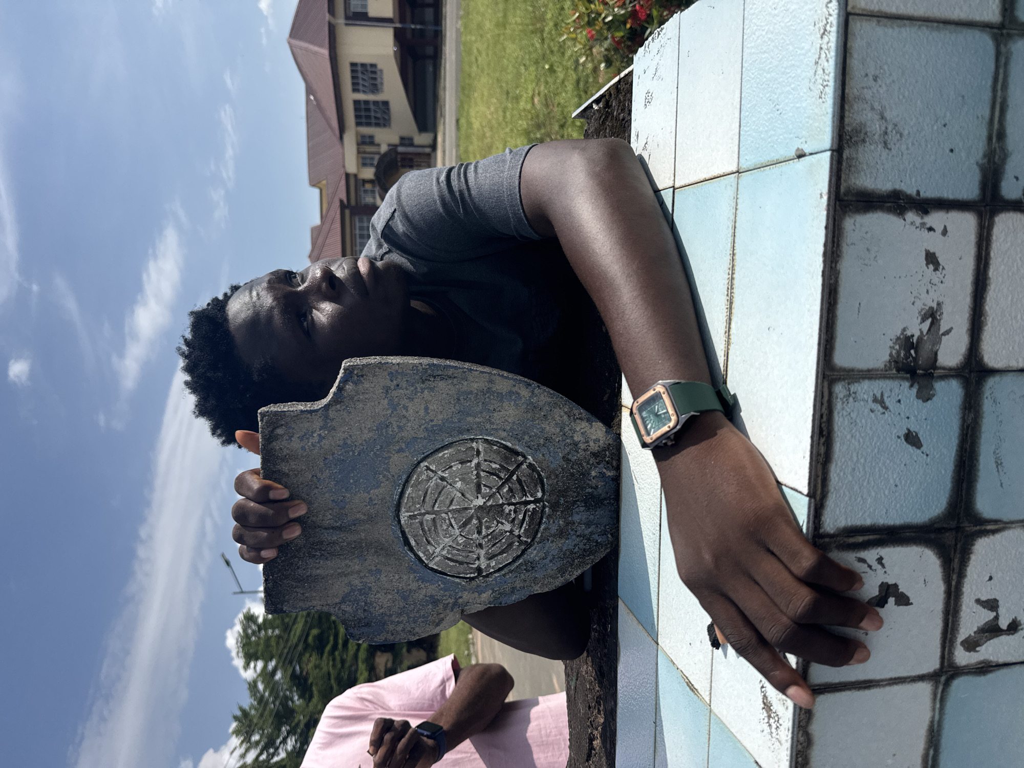 The image captures a young man outdoors on a bright, sunny day in Ihiagwa, Nigeria. The primary subject is a person with dark skin and short, dark, curly hair, wearing a grey t-shirt. He is positioned leaning on a tiled structure, looking upwards and slightly to his right. His left arm is bent, and his head rests on the structure, while his right hand holds a weathered, shield-shaped object. This object is dark grey and features a circular, intricate design resembling a compass or star emblem etched into its surface.
On his left wrist, he wears a distinctive wristwatch with a square, possibly gold-toned, bezel and a green strap. The structure he is leaning on is covered in light blue and white square tiles, some of which appear discolored or stained with dark grime. A second person, partially visible in the bottom left, wears a pink shirt and a dark smartwatch on their left wrist.
In the background, there are buildings with reddish-brown roofs, green grass, and foliage, suggesting a campus or residential area. The sky is a clear blue with sparse white clouds, indicating clear weather and daytime. The overall atmosphere is casual, and the subject's expression appears pensive or watchful. No discernible text is visible in the image.