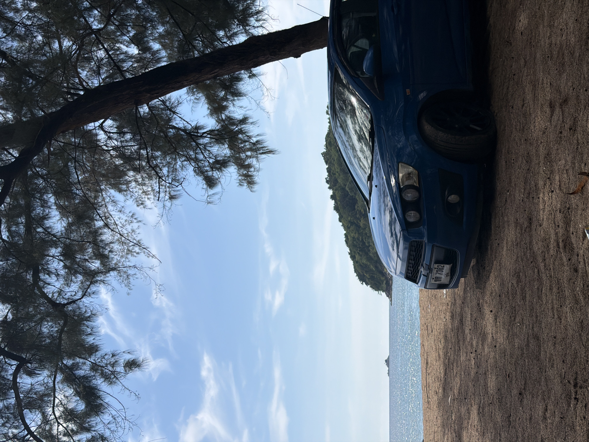 A blue car is parked on a sandy beach in Choeng Thale, Thailand. The car faces the water, and the edge of a forested area is visible on the other side of the water. A tree hangs over the car, partially obscuring the sky. The sky is light blue with wispy white clouds. The beach appears to be covered with brown sand and plant debris.