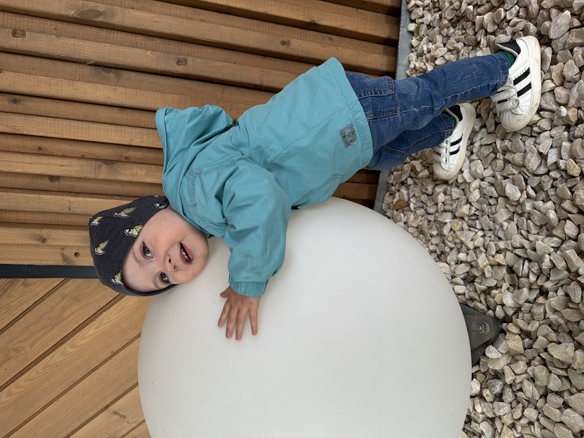 A young child wearing a blue jacket, jeans, and Adidas sneakers is lying on top of a large, white, spherical object, likely an outdoor lamp. The child is also wearing a dark gray hat with light yellow ice cream cone patterns. The child's left hand is resting on the lamp's surface, while their head is near the edge, looking at the camera. The background consists of wooden planks and a light-colored gravel surface. Based on the surrounding elements, it's plausible that the location is a park or yard in Votkinsk, Russia, where such installations might be present.