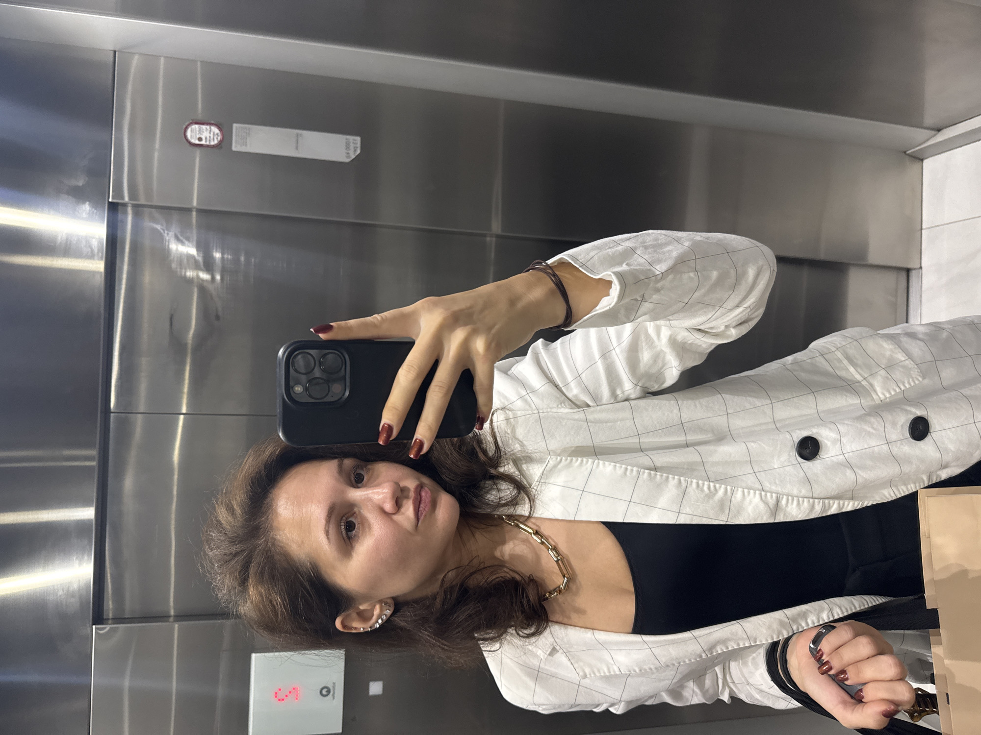 A woman takes a selfie in a stainless-steel elevator. The elevator panel shows the number "5" illuminated in red. The woman has brown hair and is wearing a white plaid blazer over a black top, along with a gold chain necklace and an ear cuff. Her fingernails are painted a dark red color. She is holding a black smartphone in her left hand, and a shopping bag is partially visible in her right hand. The elevator appears to be located in a modern building. The location is assumed to be Dubai, United Arab Emirates based on commonly available information for public elevators.