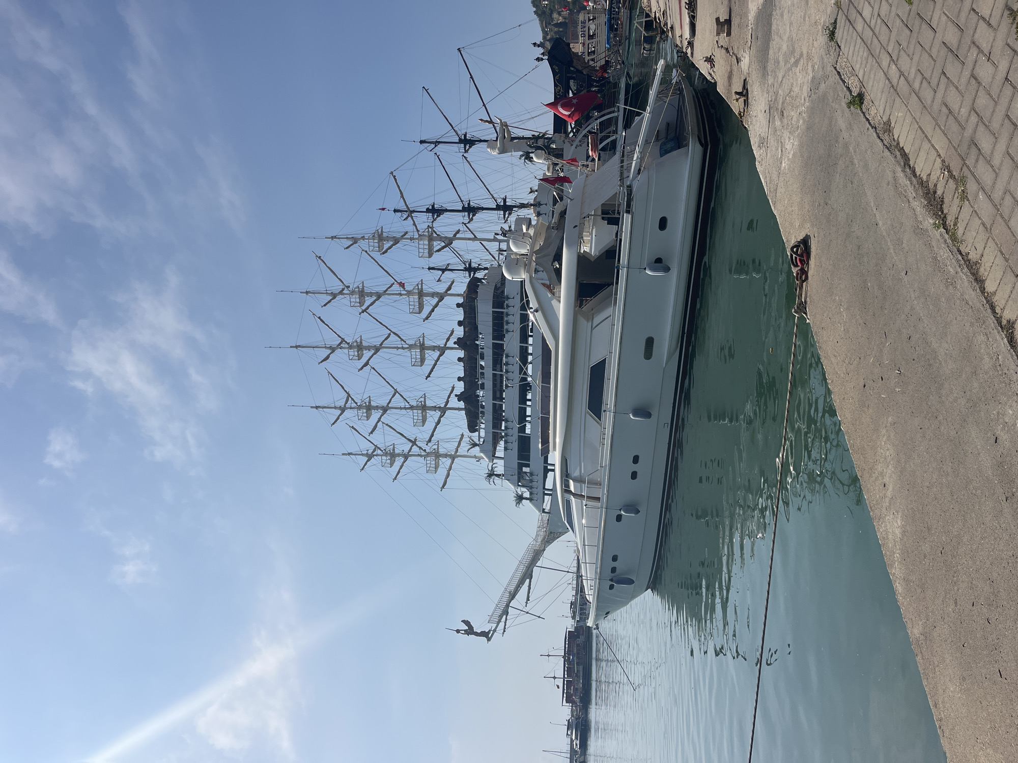 Two large vessels are prominently docked at a concrete pier under a bright, sunny sky. The image is taken from the pier, looking across calm, turquoise water towards the boats.
In the foreground, a sleek, modern white motor yacht occupies a significant portion of the frame. Its multi-deck structure features numerous windows and smooth lines. A distinctive detail is a small, dark figurine of a person in a diving pose, positioned at the very front of the yacht's bow.
Behind the white yacht, a larger, multi-masted sailing ship is moored. This vessel has a more traditional, perhaps pirate-ship inspired design, with several tall, bare masts equipped with cross spars and intricate rigging. A red Turkish flag with a white star and crescent is prominently displayed on this ship. Partially visible text on its hull or superstructure reads "SUN DIVE".
The pier itself is constructed from grey concrete, with areas of paved brickwork visible along the top edge. The water in the harbor is calm, reflecting the sky and the ships. The sky is a clear blue with sparse, wispy white clouds. Strong sunlight is evident, creating bright highlights and casting soft shadows. A noticeable sun flare is present in the lower left portion of the sky. The scene suggests a pleasant day at a busy harbor, likely a tourist destination in Alanya, Turkey.