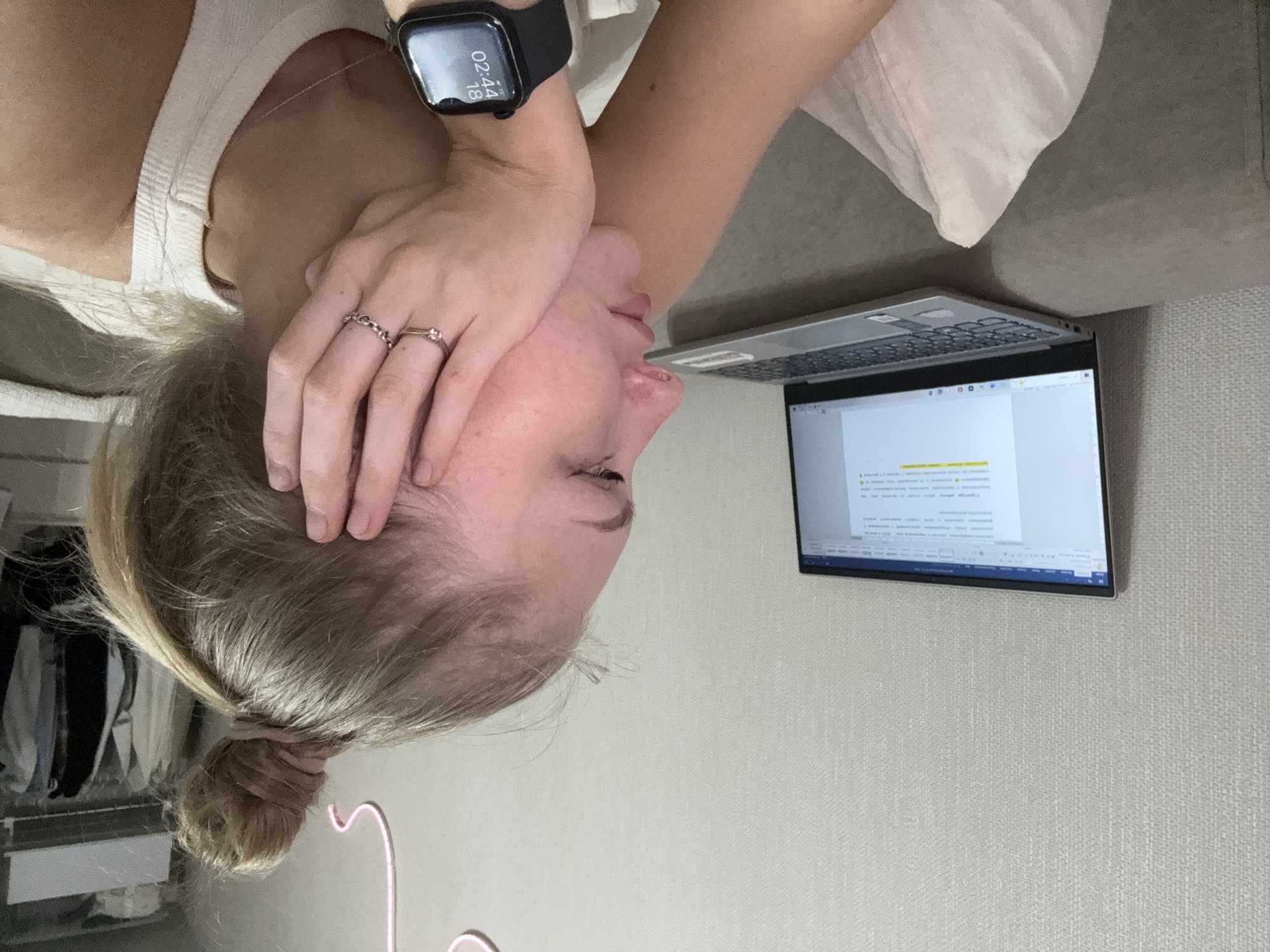A young woman with blonde hair tied up in a bun is seen from the side, resting her head on her hand. She is wearing a white tank top and has two rings on her fingers. A smartwatch on her wrist displays the time as 02:44. She is looking at a laptop, which is open and displaying a document with highlighted text. The scene appears to be indoors, possibly in a living room or bedroom, with a textured wall behind her. A glimpse of a closet with clothes is visible in the background. There is no information in the image to suggest this scene is located in the city of Perm, Russia.