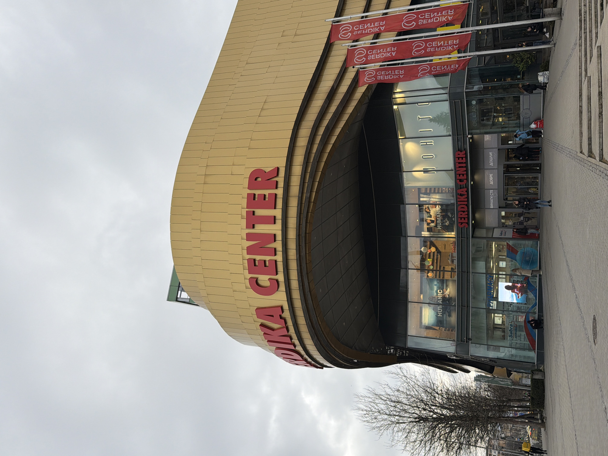 The image shows the Serdika Center, a large shopping mall in Sofia, Bulgaria. The building has a distinctive gold-colored facade with a curved shape and red lettering spelling out the center's name. Banners with the reversed words “СЕИДЕВ ZEBDIKV” are visible hanging over the entrance. There is glass frontage showing stores like "MOHITO," "HUMANIC," and "GELATIAMO". People are walking in front of the mall, with shops reflected in the mall’s glass exterior. The sky is overcast.
