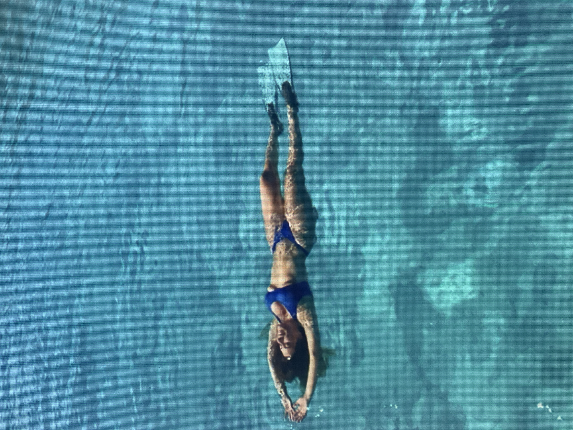 A woman wearing a blue bikini and fins is floating on her back in clear blue water. Her arms are outstretched above her head, and her legs are together. The water is calm and there are some shadows on the surface, suggesting it is sunny. The scene appears to be in an outdoor pool or natural body of water.