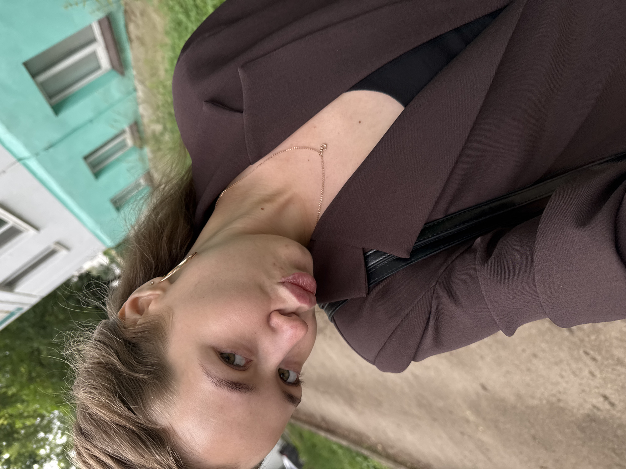 A young woman with brown hair and green eyes poses for a selfie outdoors. She is wearing a dark brown blazer over a black top and a delicate gold necklace. Her lips are pursed, and she is looking directly at the camera. The background shows a sidewalk, green grass, and a building with teal siding and white windows. This appears to be a residential area in Chaikovsky, Russia, possibly near a multi-story apartment building.