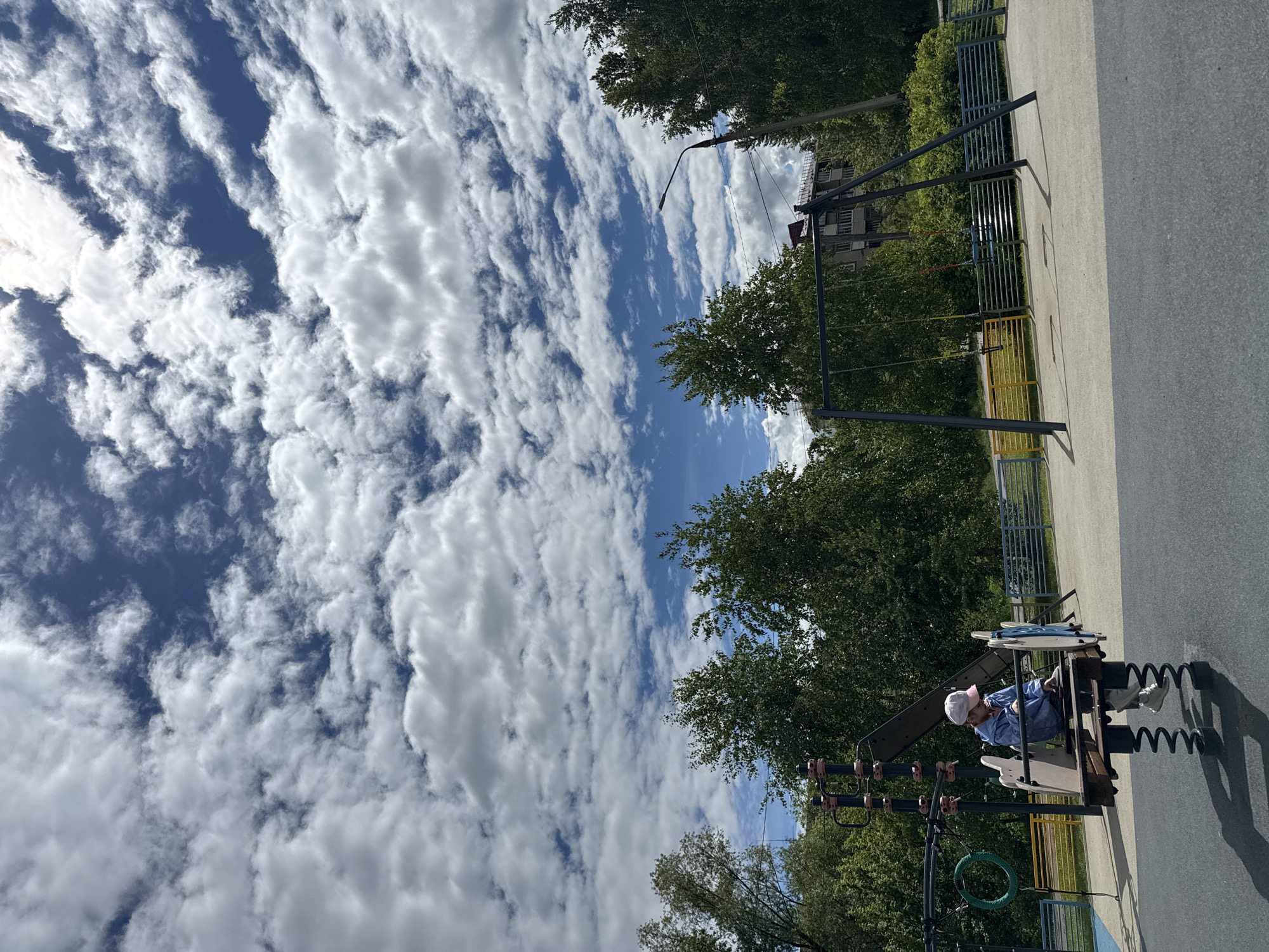 The image depicts a playground scene in Shevlyakovo, Russia. A person wearing a blue shirt, white pants, a white hat, and white shoes is sitting on a playground spring rider shaped like an airplane. Trees and a sky filled with layered, textured clouds are visible. To the right, there is a swing set. A street light and an apartment building can be seen in the background. The playground appears to be paved.