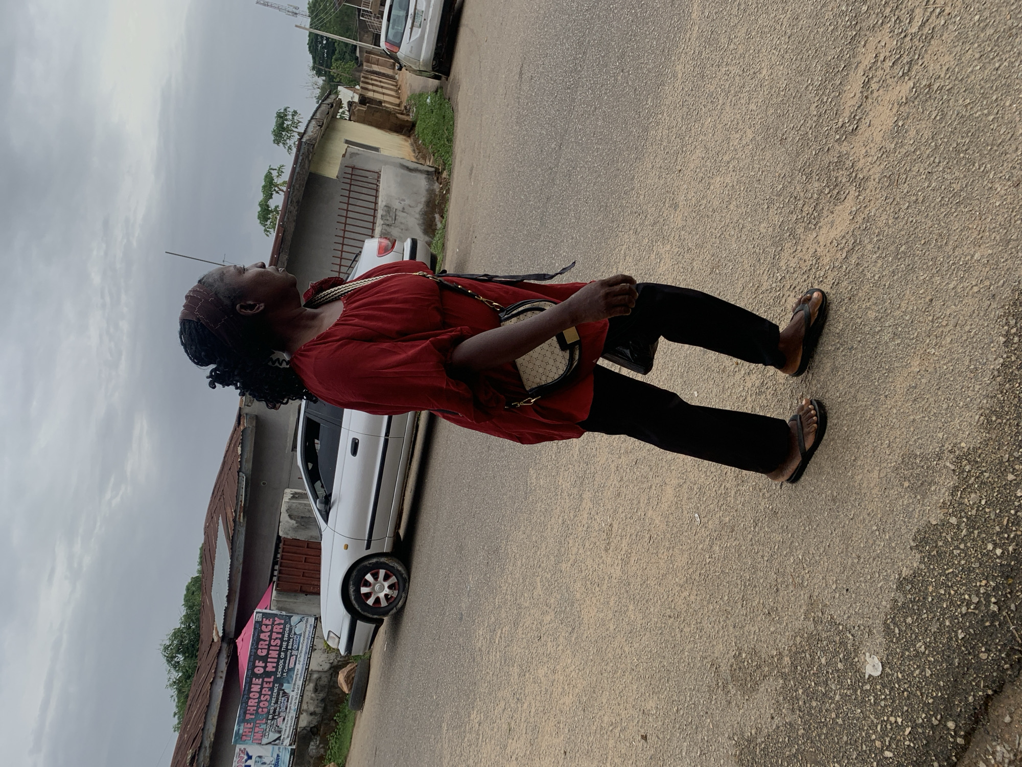 A street scene features a woman walking in an urban area. The woman is wearing a red top, black trousers, and brown sandals. She also has a handbag slung over her shoulder. She is gazing upwards. The street is paved and shows signs of wear. Along the side of the road, there are buildings with corrugated iron roofs and a white car parked nearby. A sign in front of the buildings reads "THE THRONE OF GRACE." The weather appears overcast. It is in Jos, Nigeria.