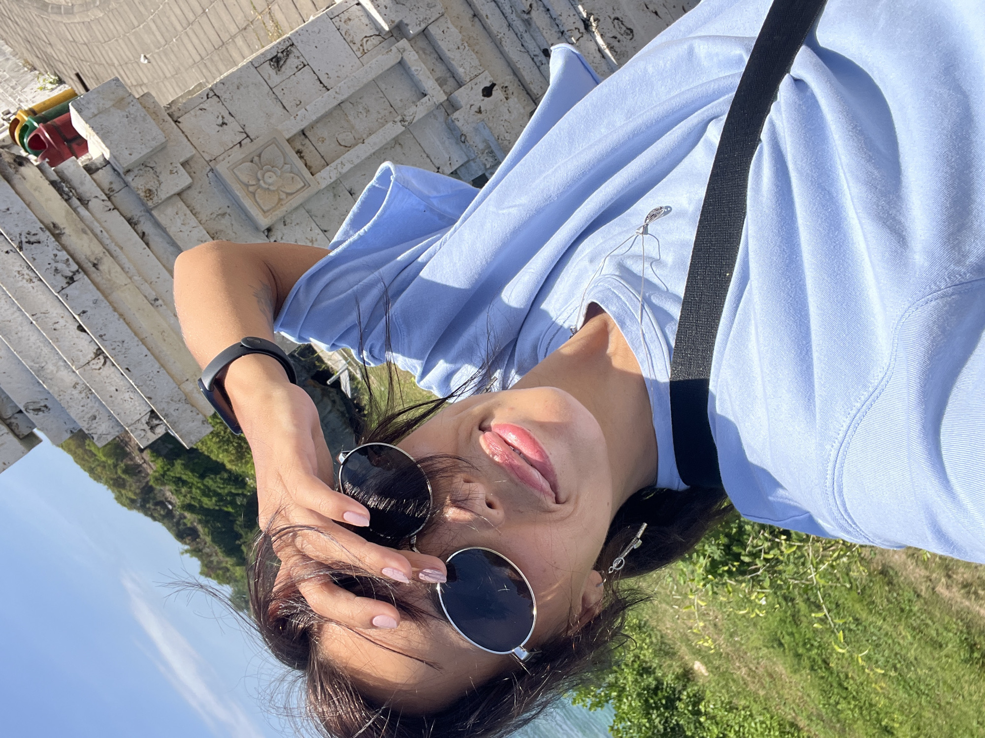 A young woman poses for a selfie in Kuta Selatan, Indonesia. She is lying on her back outside a stone structure of stacked walls, partially visible in the upper left corner. The background features bright blue sky and lush greenery.
The woman has dark hair and wears sunglasses. Her shirt is light blue and a black strap crosses her chest. She touches her hair with one hand, revealing a smartwatch on her wrist. She sticks out her tongue slightly. She has an earring in her ear, as well.