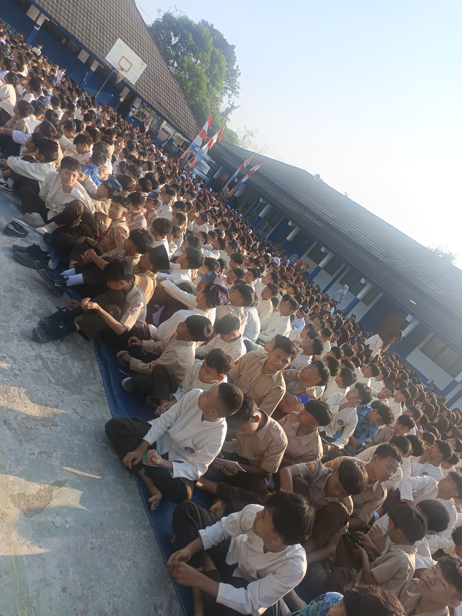 The image depicts a large group of students, likely elementary or middle school age, seated on a blue mat on a concrete surface, outdoors. The students are wearing uniforms, with many in light-colored shirts and dark pants, and some in khaki shirts and dark pants. They are arranged in rows and appear to be attending an event or assembly, as many are looking in the same direction.
In the background, a long, multi-story building with a dark tiled roof is visible. The building has blue and white striped accents along its facade, and a basketball hoop is mounted on one of its walls. Trees are present behind the building, and the sky is bright and clear, suggesting a sunny day. Two Indonesian flags are visible on poles near the building. The overall scene suggests a school setting in Cianjur, Indonesia.