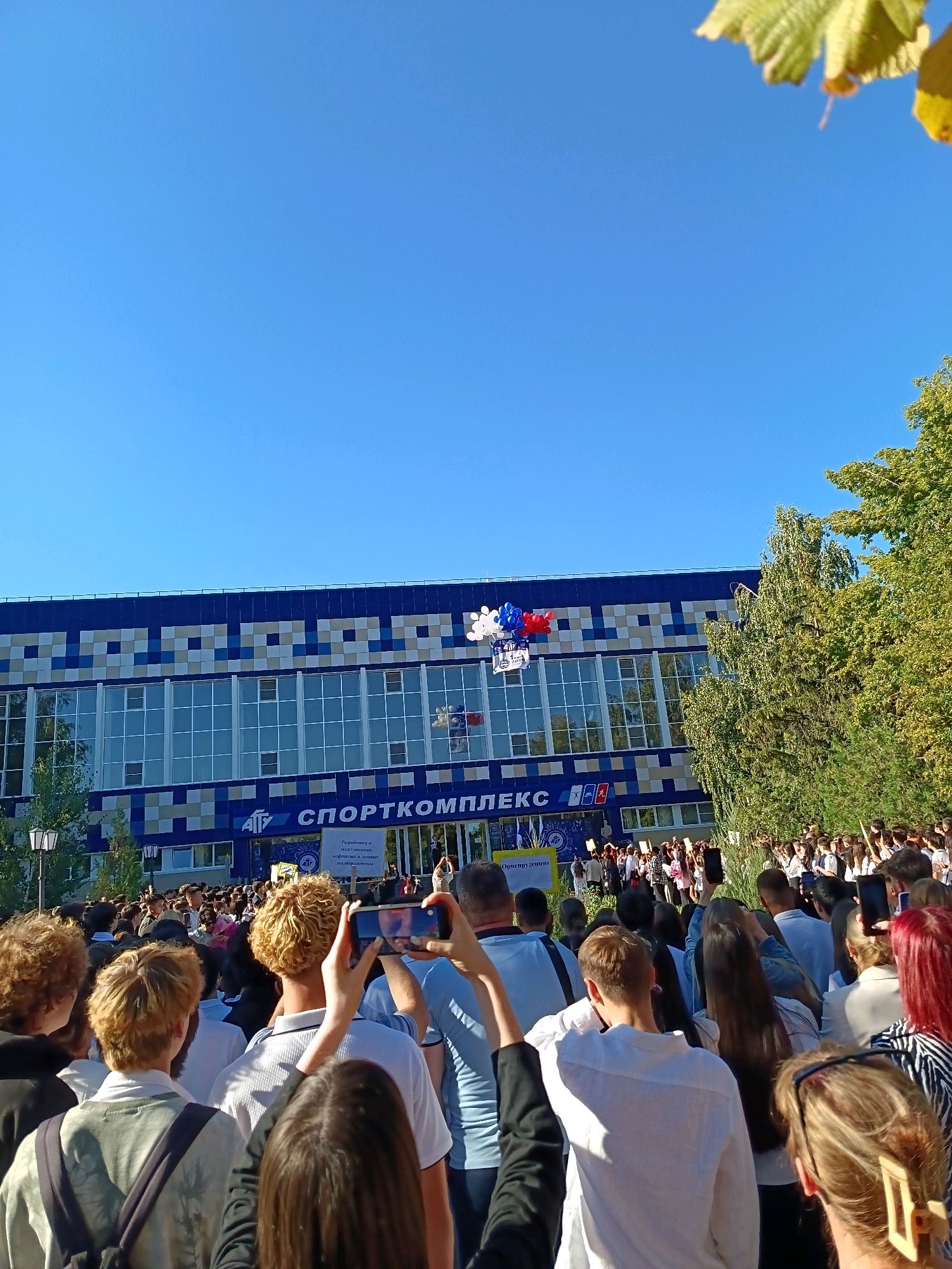 A wide shot captures a crowd of people gathered outdoors in front of a large, blue and white tiled building with "СПОРТКОМПЛЕКС" (SPORTS COMPLEX) written in large letters above the entrance. The building has numerous windows, and a banner with Cyrillic text is visible near the entrance. A cluster of red, white, and blue balloons floats in the air near the building. Many people in the crowd are holding up their mobile phones, some to take pictures or videos. The sky is clear and blue, and the sun is shining, casting shadows. Trees with green leaves are visible on the right side of the frame, suggesting an outdoor setting in a city. The overall scene appears to be an event or gathering.