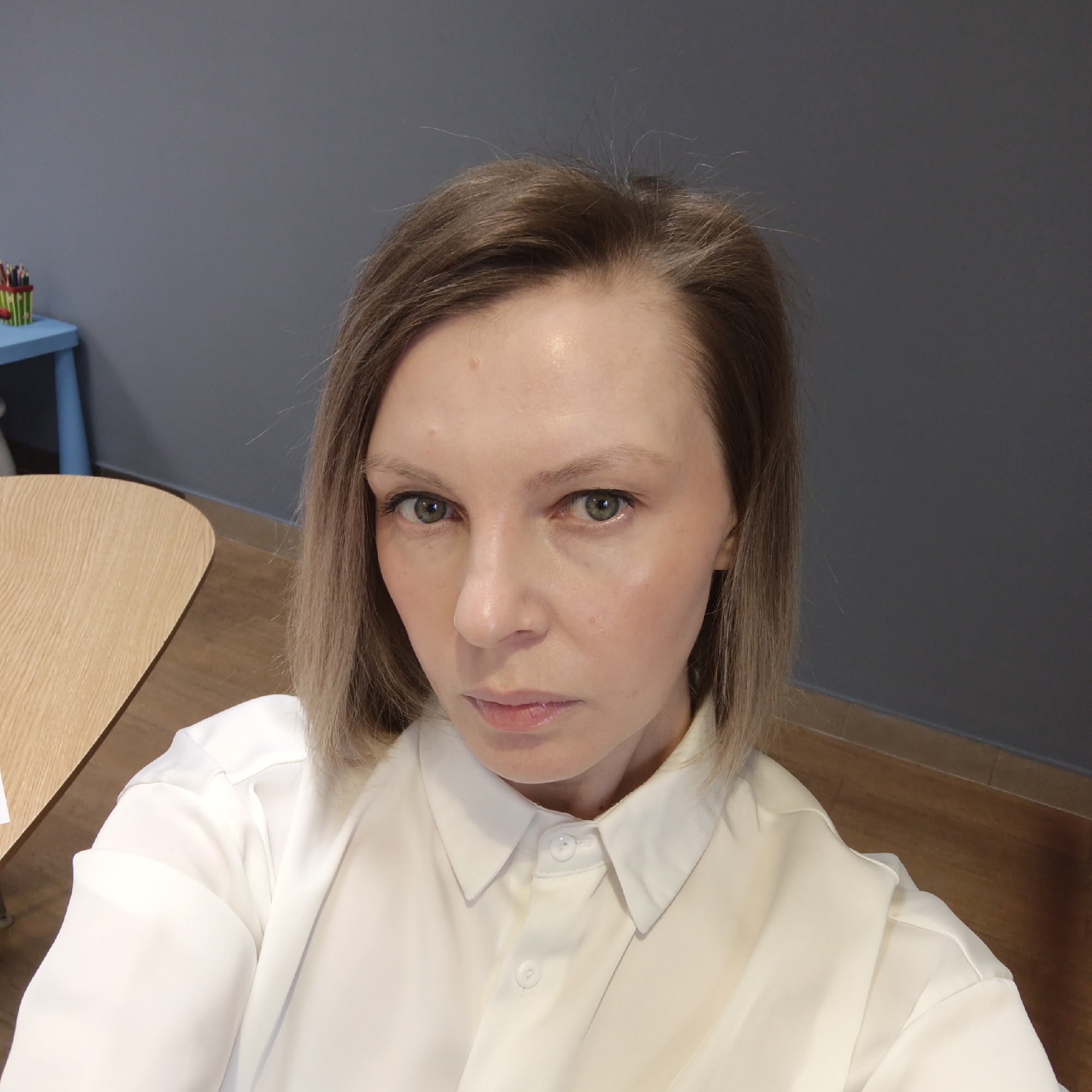 A medium shot shows a Caucasian female with short blonde/light brown hair, wearing a white collared shirt. She is looking directly at the camera with a neutral expression. Her eyes are green. The background is a plain gray wall. In the lower left, a portion of a light wood table is visible. In the upper left corner, there is a small blue table or shelf with a container of writing utensils on it. The flooring is a light brown wood grain. The location is likely an indoor space in Chaikovsky, Russia.