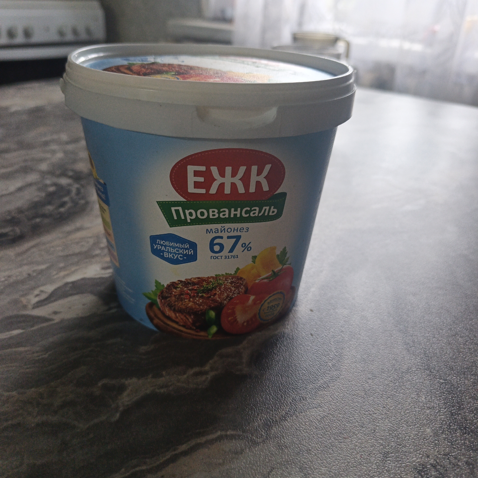 A close-up, eye-level, slightly angled shot shows a light blue plastic tub of mayonnaise on a dark gray speckled countertop. The tub has a white lid with a ribbed edge. The label on the tub features a red oval with white Cyrillic lettering that reads "ЕЖК". Below this is a green banner with white Cyrillic text that says "Провансаль". Further down, the label indicates "майонез" and "67%" in large font, with "ГОСТ 31761" below. A section of the label on the left, in a blue badge, reads "ЛЮБИМЫЙ УРАЛЬСКИЙ -ВКУС-". The bottom of the label displays a photographic image of a steak, a half tomato, a slice of lemon, and some green herbs or peas. The background is out of focus, showing what appears to be a kitchen appliance and a window with sheer curtains. The image is well-lit, with the primary subject in sharp focus. There is no indication of the specific location within the city of Юшала, Russia, beyond what can be inferred from the product's branding.