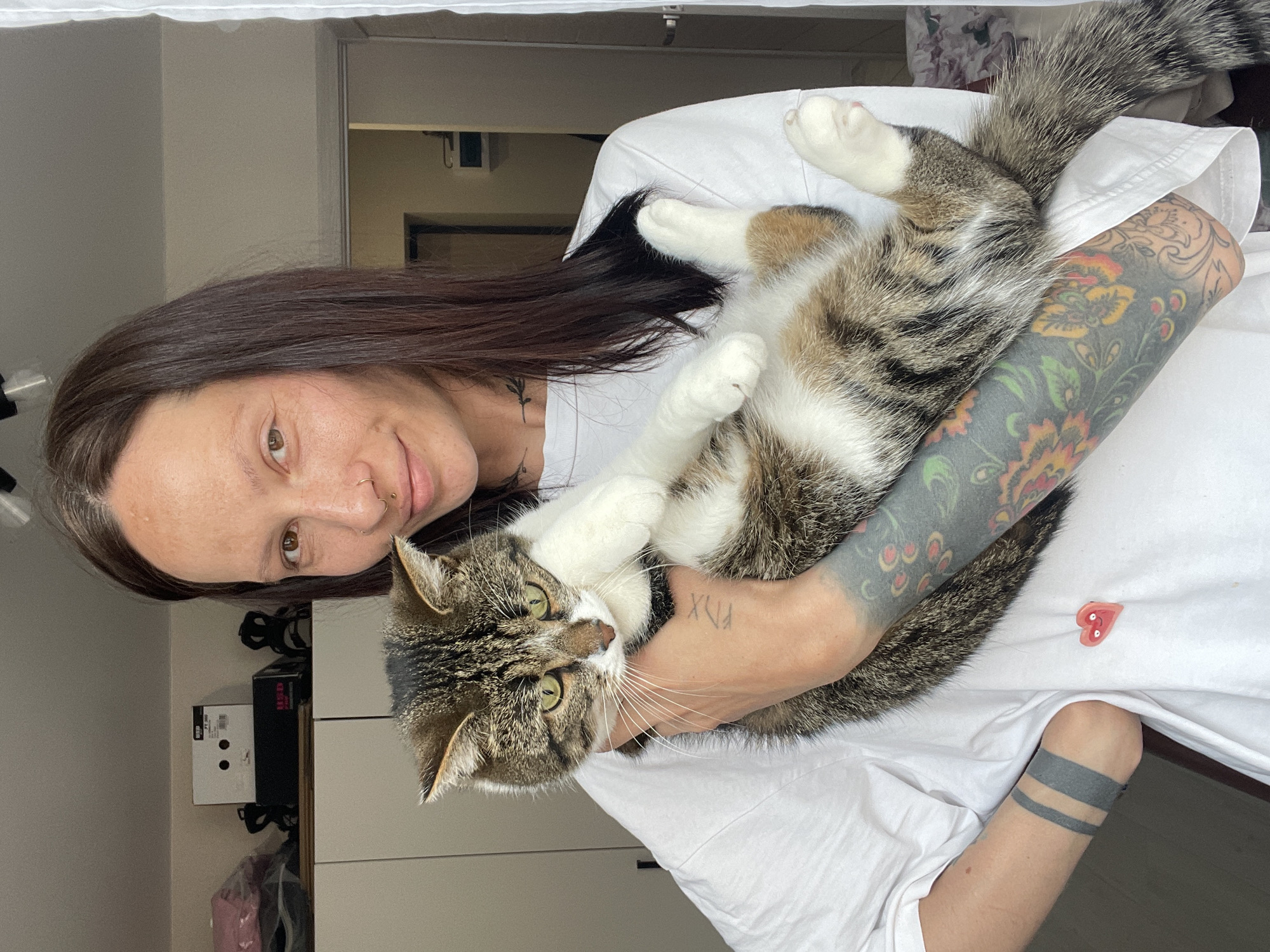 A woman is holding a cat in her arms. The woman has dark hair, a nose ring, and tattoos on her arms and neck. The cat is a tabby with white paws. The background is a room with white walls and a window. There is a black box on a shelf behind the woman. The scene is in a residential interior, details insufficient to establish location in Pervomaysky, Russia.
