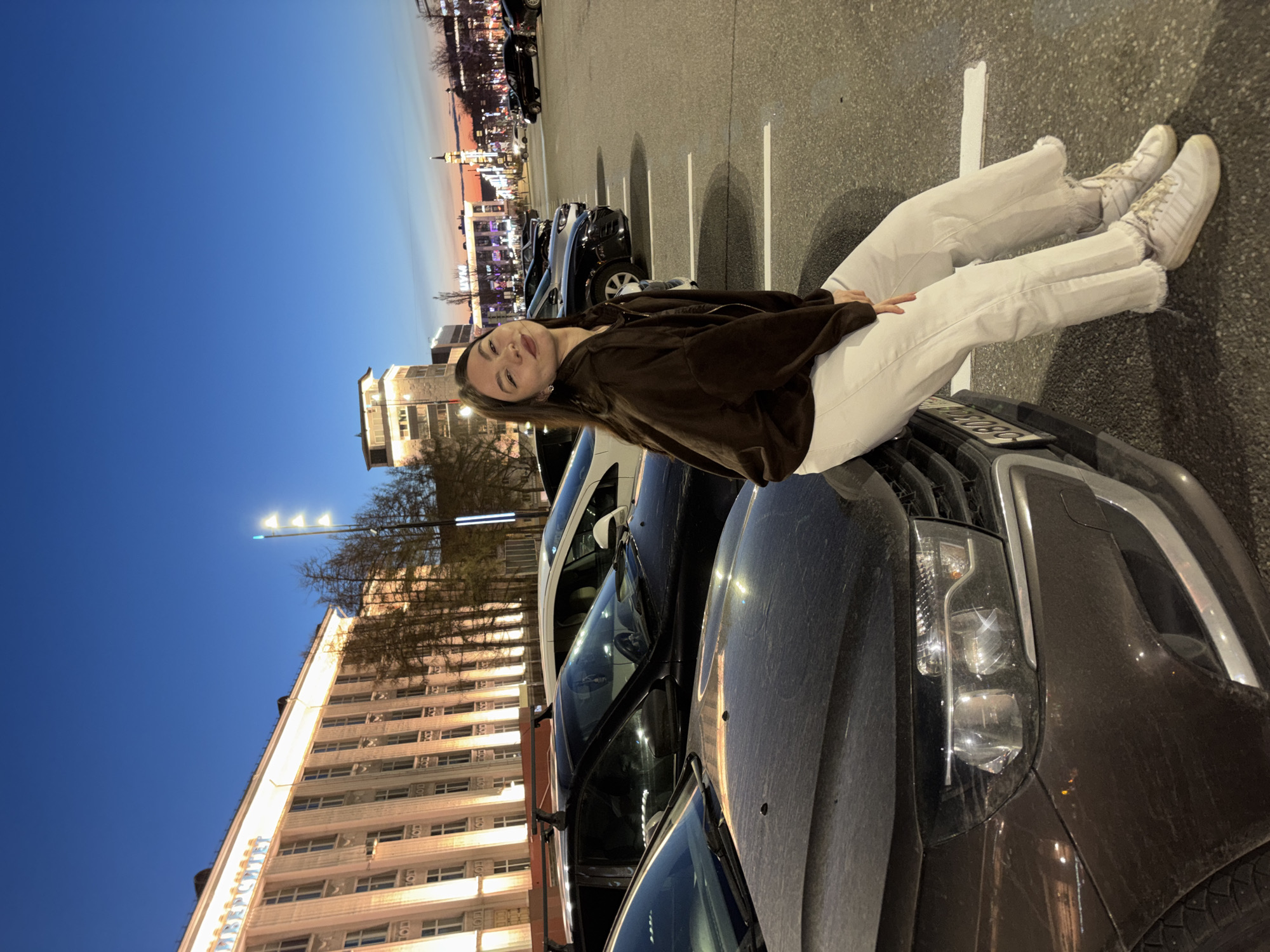 A young woman with long brown hair, wearing a brown jacket, white pants, and white sneakers, is lying on the hood of a dark-colored car in a parking lot at dusk. She is looking at the camera. Behind the car is another car. In the background, there is a tall, light-colored building with many windows and streetlights. Further in the background, other buildings and lights can be seen, suggesting an urban environment. Based on the architecture and the color temperature of the lights, it appears to be the city of Perm, Russia.