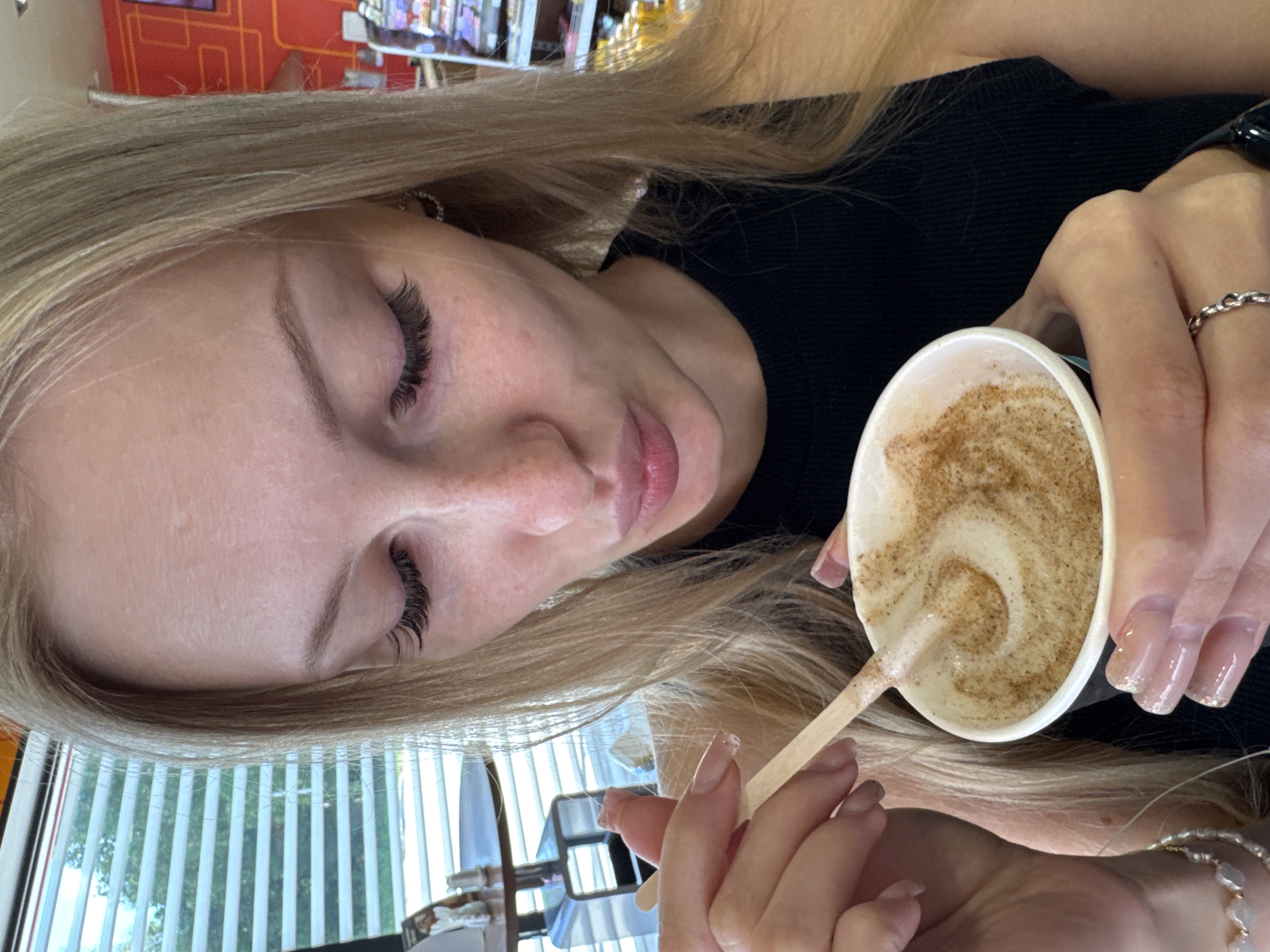 A young Caucasian woman with long blonde hair, wearing a black tank top, is holding a paper cup filled with a foamy beverage topped with cinnamon. She is using a wooden stirrer to mix the drink and is leaning in as if to taste it. The woman has long, dark eyelashes and light pink lips. She is wearing several rings on her fingers and a pearl bracelet on her left wrist. In the background, there are horizontal blinds on a window, through which trees are visible. A table with salt and pepper shakers and a small glass dispenser is also visible near the window. To the right, there are shelves stocked with products, suggesting a commercial setting, possibly a cafe or convenience store. The overall scene is brightly lit. The location is identified as Marchugi, Russia.