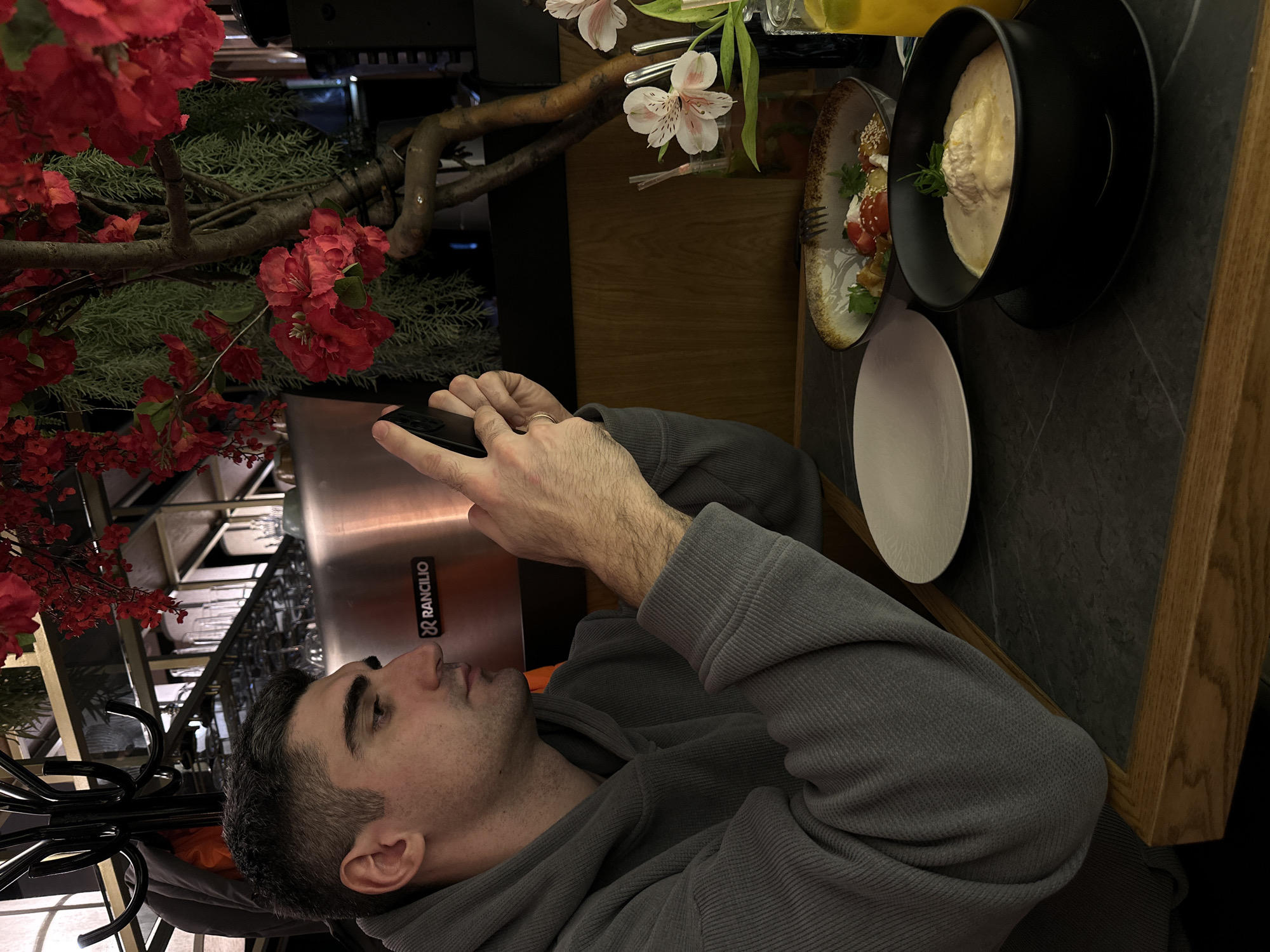 A man sits at a table in what appears to be a restaurant. He is looking at a smartphone in his hands. On the table, there are food items: a bowl of soup in a black bowl, a plate of salad, a plain white plate, and a drink in a glass. There is a floral arrangement with red and white blossoms near the table. The location is in the city of Ovoshchnoy, Russia.