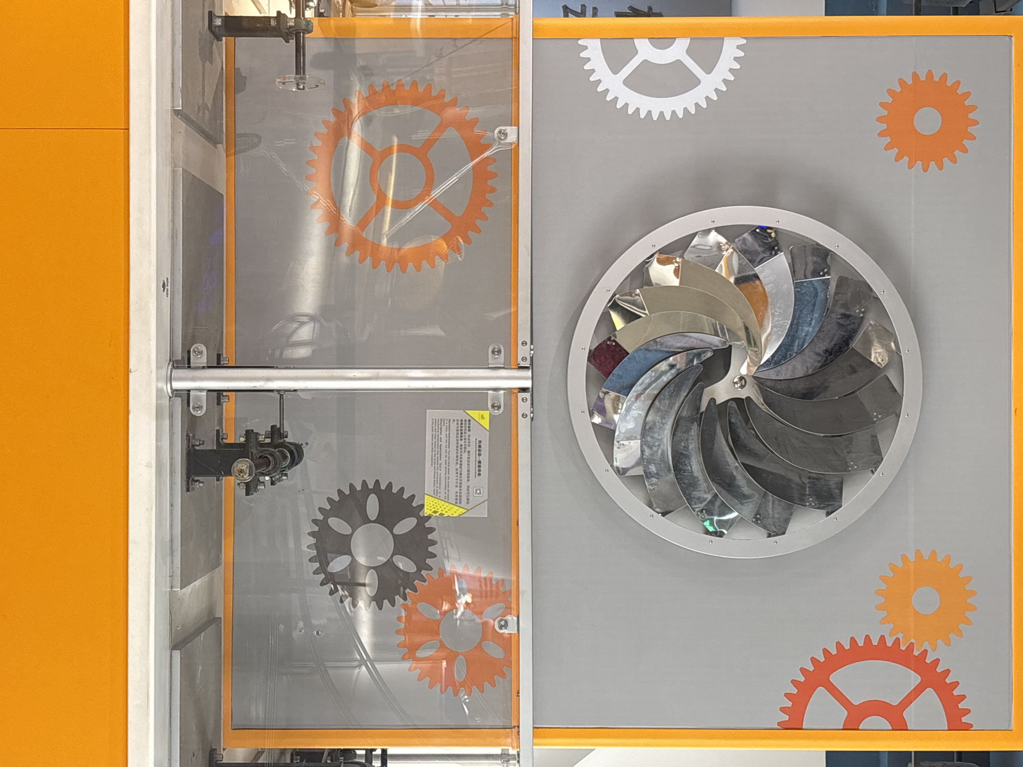 The image shows an indoor exhibit, possibly related to science or technology, in Jiangjin, China. On the left, there are two transparent panels with orange frames. The upper panel features an orange gear graphic, while the lower panel has both a black and an orange gear graphic. A silver bar runs horizontally across the center of these panels, and a black mechanical device is attached to the upper panel. To the right of these panels is a gray background with a large, circular metal sculpture resembling a turbine or fan. Several gear graphics in white and orange are scattered around the sculpture. The colors orange, silver, black, and white dominate the scene. A small sign in Chinese is visible on the lower left panel.