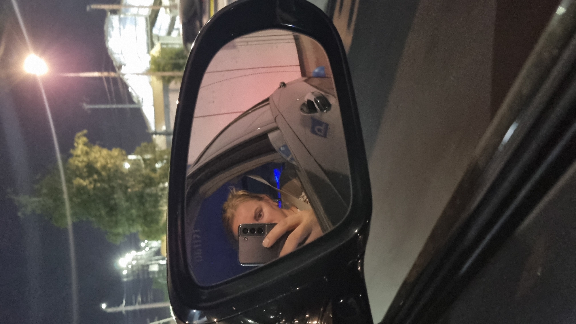 A nighttime selfie is taken in a car's side mirror, reflecting a young woman holding a phone to capture her own image. She is looking directly at the camera, her blonde hair styled casually. She wears a light-colored top with a patterned scarf or necklace. The car's interior is dimly lit, with a blue glow emanating from somewhere in the background.
Outside the car, streetlights illuminate the scene. A tall tree with dense green foliage stands prominently in the background, its branches reaching towards the night sky. Beyond the tree, a modern building with a metallic facade and a distinct geometric roofline is visible. Streetlights with bright bulbs are dotted across the landscape, casting a warm glow. The overall setting suggests an urban environment in Reutov, Russia. The reflection in the mirror also shows a part of the car's exterior, including a door handle and a parking symbol. The road surface is visible in the foreground, appearing dark and wet.