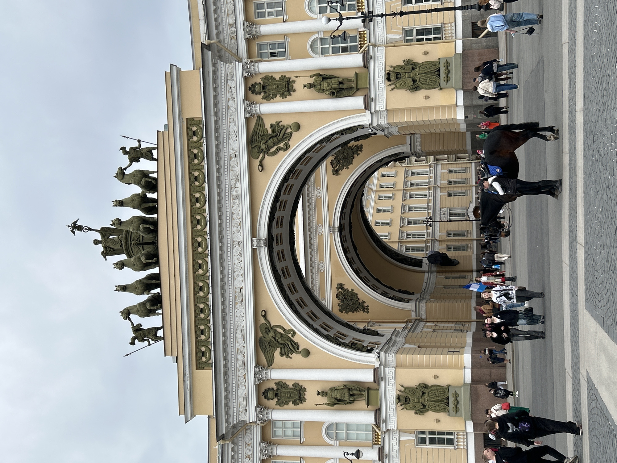 The image depicts the General Staff Building in Saint Petersburg, Russia. The building is a large, yellow structure with a prominent triumphal arch. The arch is adorned with sculptures and decorative elements, including horses and figures on top. Below, a group of people are seen walking through the archway and standing around. A horse and rider are also visible in the foreground. The ground is paved, and the sky is overcast.