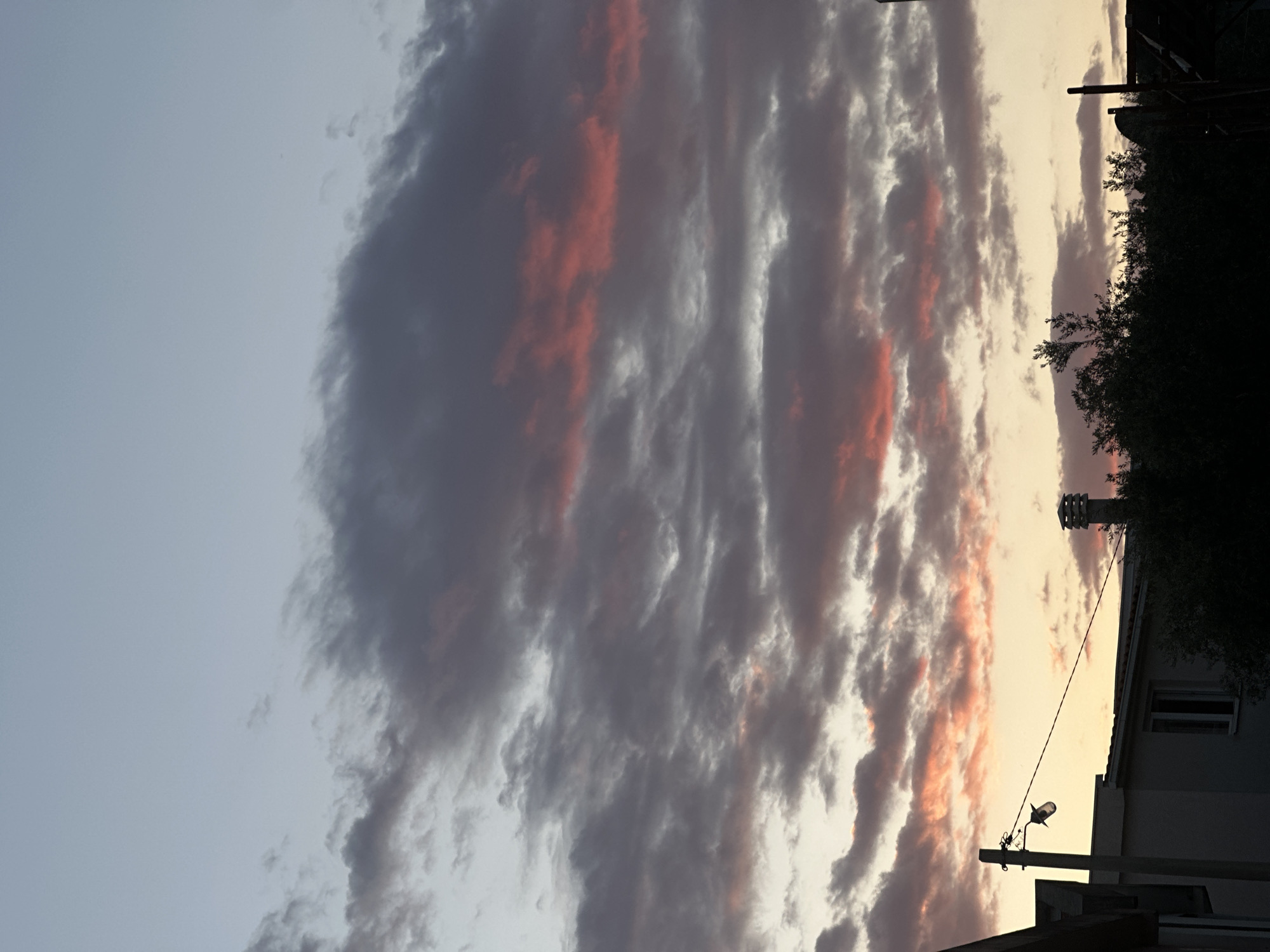 The image is a vertical shot of a sunset sky in Bar, Montenegro. The sky is predominantly gray with pink-orange hues highlighting the cloud formations. The scene includes the silhouette of a building, trees, and a utility pole with electrical wires against the bright horizon. The building has a window, chimney, and birdhouse. The scene is taken during twilight.