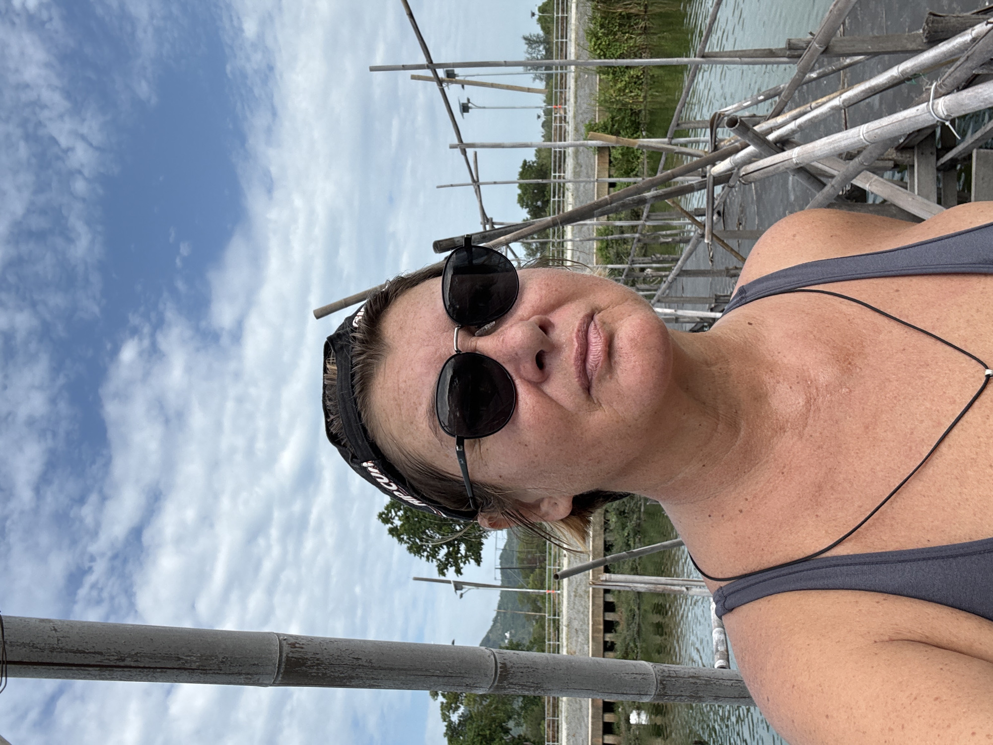 A woman takes a selfie outdoors during daylight. She is wearing a gray tank top, sunglasses, and a black headband with white text. In the background, there is a partially obscured view of a landscape, including bamboo structures, water, vegetation, and distant hills. The image was taken in Baan Chaweng, Thailand.