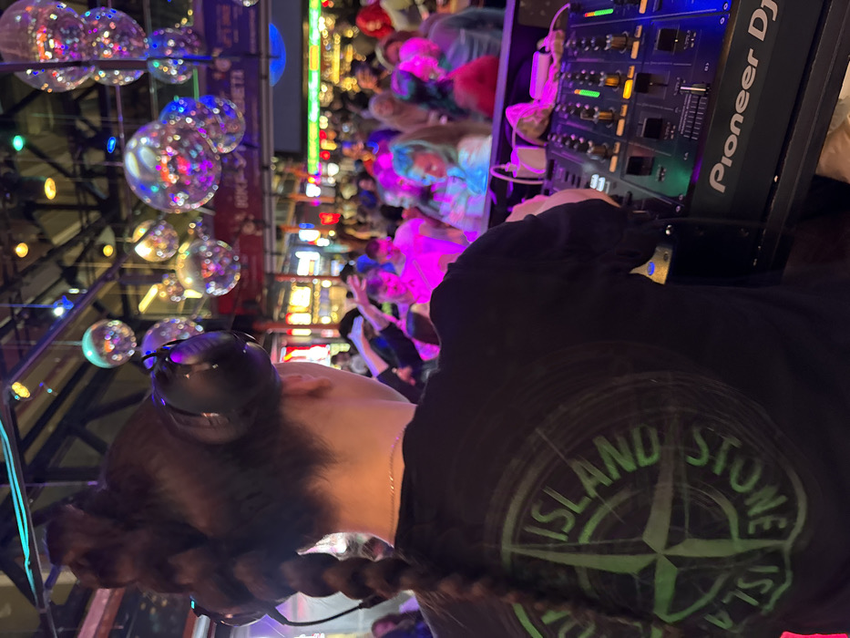 An individual, viewed from behind, is operating a Pioneer DJ mixer in a crowded indoor venue. The subject wears a black long-sleeved shirt with a prominent green "STONE ISLAND" compass logo on the back, and black over-ear headphones. Dark hair is styled with a long braid visible down the back. The DJ mixer features illuminated controls and the "Pioneer DJ" brand name.
The background reveals a large gathering of people, out of focus, illuminated by colored lights, predominantly pink. An industrial-style ceiling holds numerous reflective disco balls. Distant commercial signage in Cyrillic is partially visible, including the word "ПЛАНЕТА" (PLANETA). The scene depicts an active entertainment or food hall environment.
The location is identified as the **Depo Food Mall (Депо Лесная)** on Lesnaya Street in Moscow, Russia.