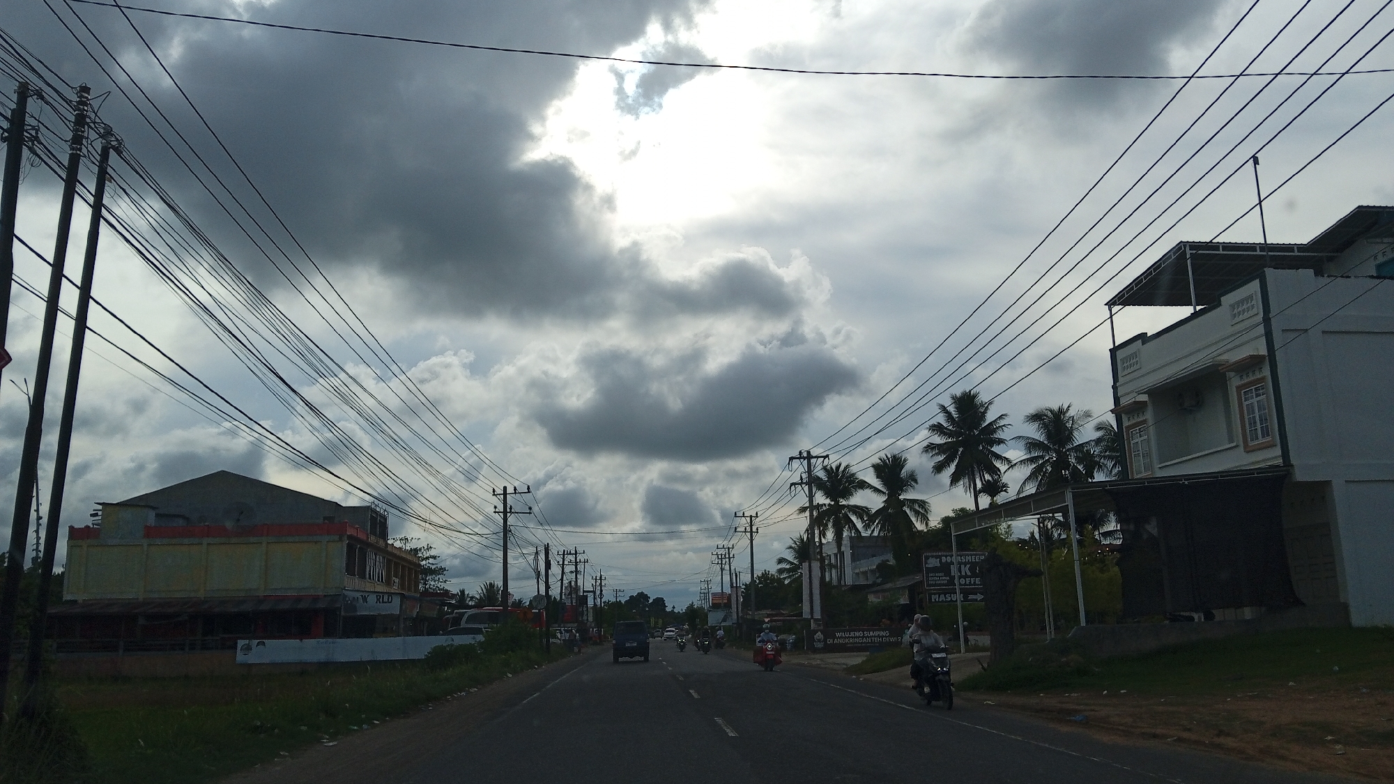 This is a street-level, first-person perspective photo of a road in Payameuneng, Indonesia. The sky is overcast with dramatic dark clouds, and the sun is attempting to break through. Utility poles with a dense network of electrical wires dominate the foreground and extend into the distance.
On the right side of the road, a modern, multi-story building with a light green facade is visible, featuring windows with decorative trim. A covered outdoor area is attached to the front of this building. Further down the road on the right, there are signs, one of which reads "DOORSLOOP COFFEE." Palm trees are interspersed throughout the scene.
On the left side of the road, a long, low-rise building with a yellowish facade and red trim is present, with what appears to be a corrugated metal roof covering an open-air area at the front.
The road itself is asphalt and appears to be a two-lane road with vehicles traveling in both directions. Several motorcycles and at least one car are visible on the road, indicating moderate traffic. One person is riding a motorcycle on the right side of the road. The environment appears to be a semi-urban or suburban area.