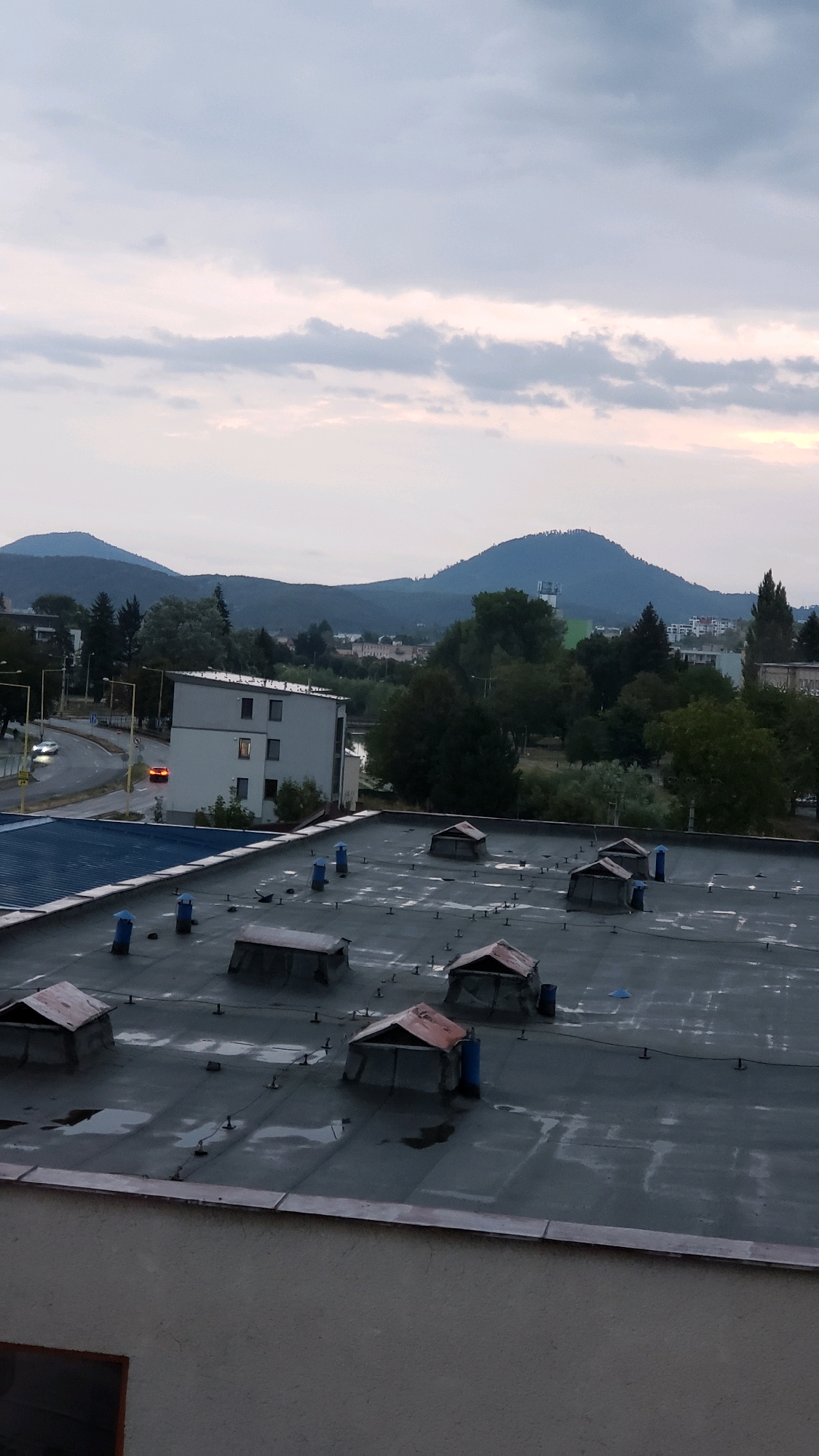 This is a vertical, medium-distance, outdoor, eye-level shot taken on a cloudy day. The scene is dominated by a flat, grey, slightly wet rooftop in the foreground, littered with several small, dark-colored ventilation structures with reddish-brown angled tops. Some blue cylindrical objects are also visible on the rooftop, along with a single blue triangular object. In the middle ground, a modern, three-story white building with dark windows and one lit window is visible. To the left of this building, a curving road with streetlights and cars can be seen. Further in the background, rolling hills and mountains are covered in trees, with some buildings scattered amongst them. The sky is overcast with soft grey and white clouds, with hints of pink and yellow light suggesting either sunrise or sunset. The overall impression is of a city, likely Prešov, Slovakia, viewed from a rooftop.