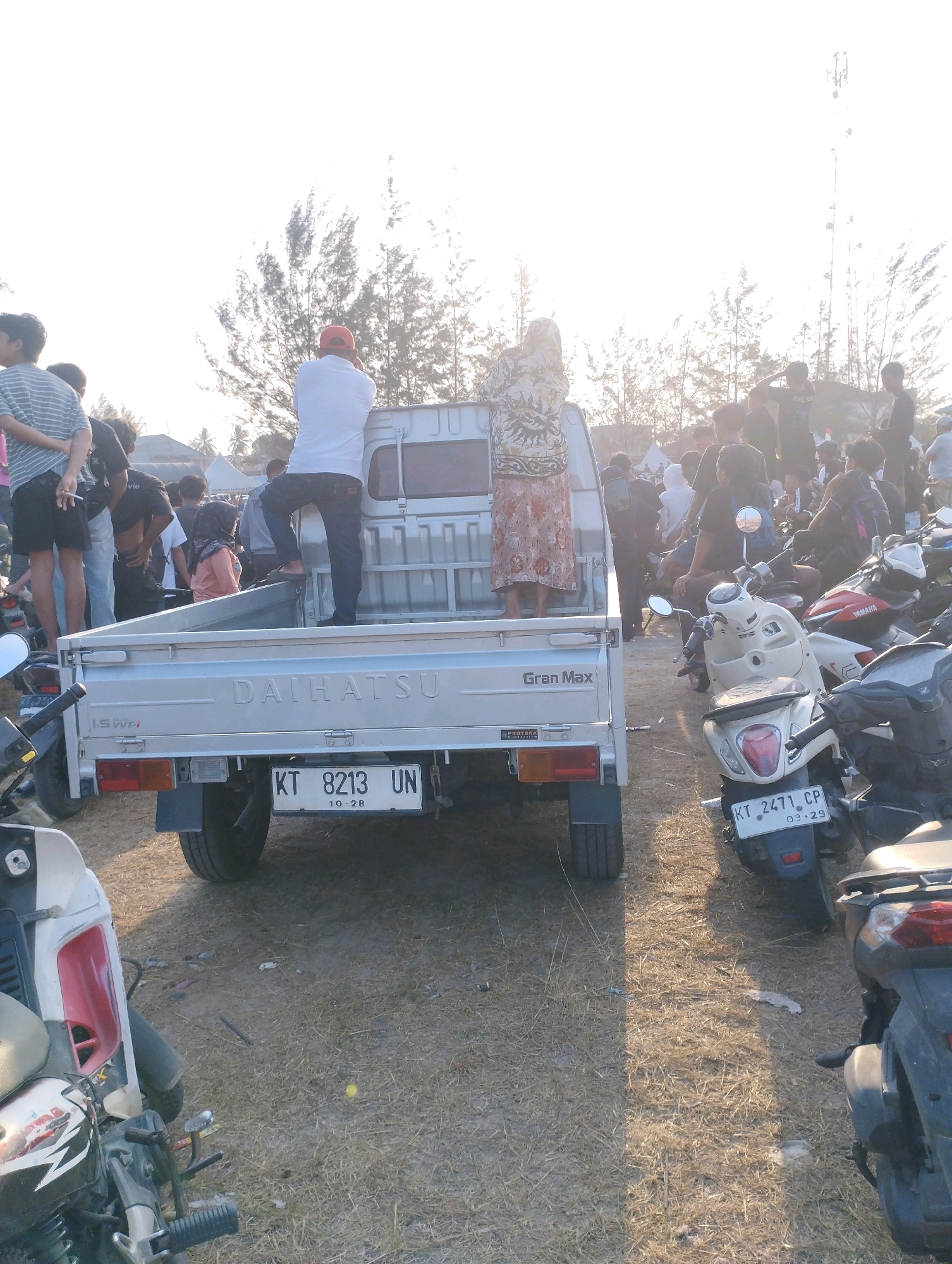 The image depicts a gathering of people and several motorcycles parked on a dirt and dry grass ground. A silver Daihatsu Gran Max pickup truck is positioned prominently in the foreground, with its tailgate open and a license plate reading "KT 8213 UN". Two individuals are standing on the tailgate of the truck; one man wearing a white t-shirt and red cap is facing away from the camera, while a woman in a patterned dress is also facing away. Other individuals are visible in the background, standing around and near motorcycles. The environment appears to be an outdoor space with sparse trees and a bright, hazy sky, possibly suggesting a warm climate or late afternoon light. The scene suggests a casual social gathering or event.