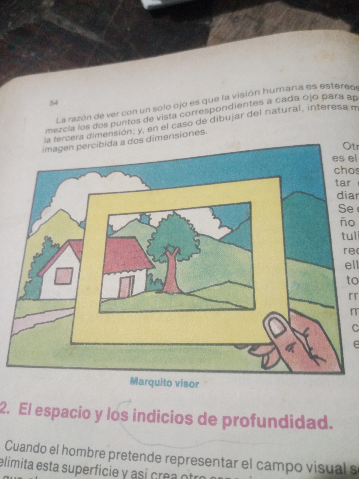This is a close-up, eye-level, indoor photograph of an open book, with the focus on a page containing text and a colored illustration. The page is aged and slightly yellowed.
The illustration depicts a simplified, colorful landscape scene. In the foreground, a hand is holding a yellow rectangular frame, as if looking through a viewfinder. The scene within the frame includes a house with a red roof, a tree, green rolling hills, and a light blue sky with white clouds.
To the left of the illustration, there is text in Spanish. A small number "54" appears in the upper left corner of the text block. The text discusses human vision and depth perception. Below the illustration, there is a caption in Spanish that reads "Marquito visor". Further below this, a larger heading in pink font reads "2. El espacio y los indicios de profundidad." ("2. Space and the indications of depth."). Below this heading, there is more Spanish text.
To the right of the illustration, there is more Spanish text, with some words partially visible.
The photograph's lighting appears to be from a single source, creating some subtle shadows. The background behind the book is dark and out of focus.
There is no information within the image that provides any geographical context to Santa Ana, Venezuela.