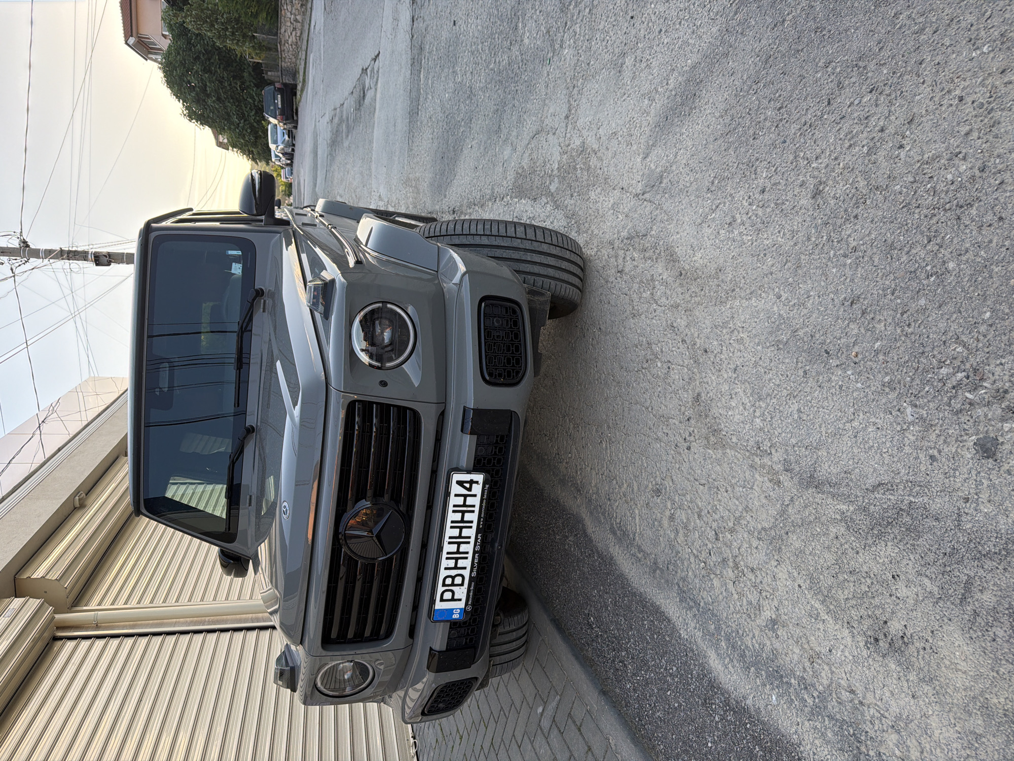 A grey Mercedes-Benz G-Class SUV is parked on a grey asphalt road. The front of the car is visible, showcasing its grill, headlights, and Bulgarian license plate, "PBHHHHH4". The vehicle appears to be in Asenovgrad, Bulgaria. A commercial building is to the side. Other vehicles are on the road in the background.