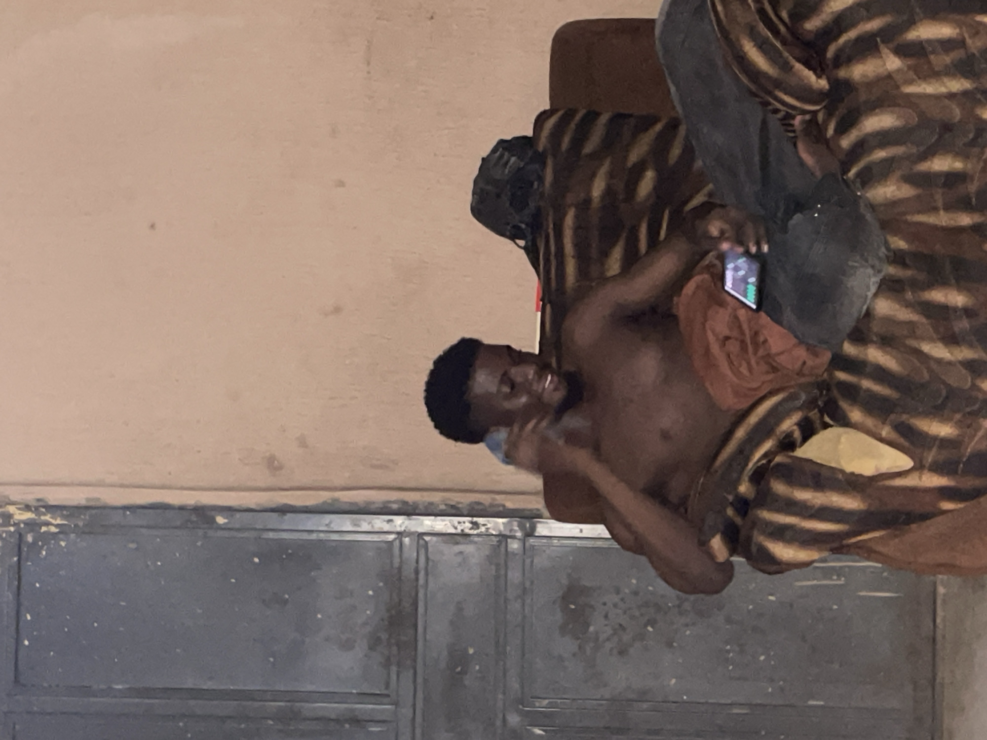 A shirtless Black male is seated on a couch with a zebra-striped patterned upholstery, indoors. He is holding a smartphone in his right hand and appears to be looking at it. A brown cloth is draped over his lap and the couch. To his right, a black backpack is visible on the back of the couch. The background features a textured, light-colored wall and a dark metal door on the left. The setting is likely a residential area in Lokoja, Nigeria, given the typical building materials and aesthetic.
