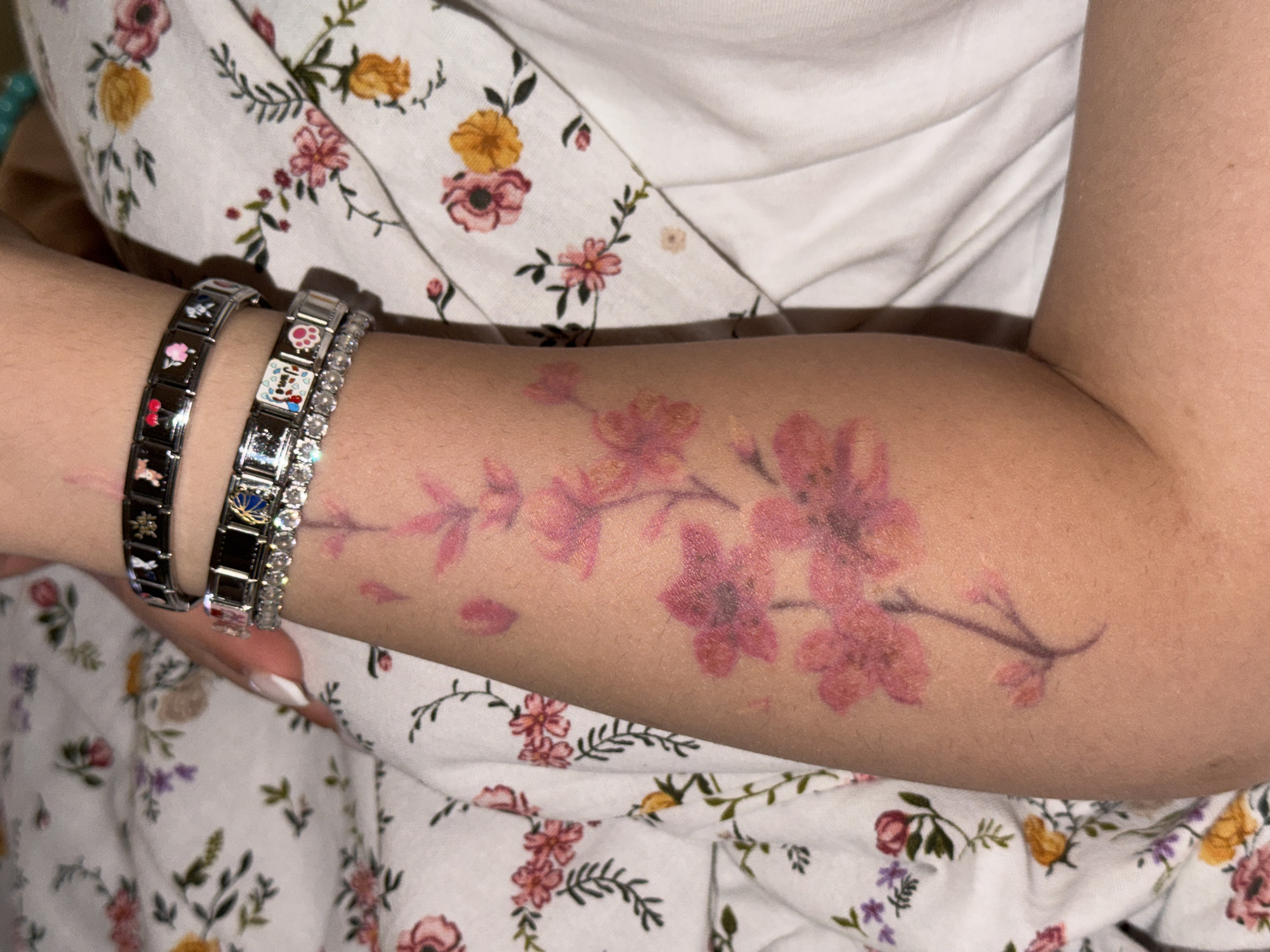 The image presents a close-up view of a person's arm, adorned with jewelry and a tattoo, resting against a floral fabric background. The arm, with a fair skin tone, is positioned diagonally across the frame.
On the wrist, three bracelets are visible. On the left, there is a silver-toned Italian charm bracelet featuring black enamel links with various colorful designs, including a red cherry and a pink flower. Adjacent to it, another similar charm bracelet displays designs such as a paw print, a golden crown with blue accents, and a motif of what appears to be a character or animal on a white background. Nestled between these charm bracelets and slightly to the right is a sparkling silver-toned tennis bracelet set with numerous clear stones.
Further up the forearm, a tattoo depicts a flowing branch of cherry blossoms. The flowers are rendered in shades of faded pink and reddish-purple, with some individual petals appearing to fall, suggesting a delicate and artistic design.
The background consists of a white fabric, likely a piece of clothing, patterned with a dense scattering of small flowers in various hues including pink, yellow, and purple, along with green leaves and stems. The lighting appears to be soft and even, typical of an indoor setting. No discernible text is present in the image.