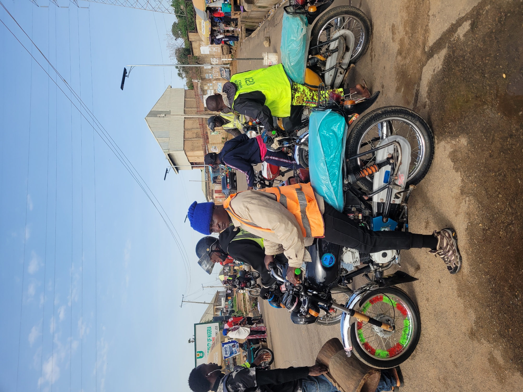 The image captures a street scene in Gura Top, Nigeria, with several individuals gathered around motorcycles. In the foreground, a person wearing an orange safety vest and a blue beanie is seated on a black motorcycle. To their left, another person in a black shirt and jeans stands, holding a large wooden cylindrical object. Behind them, other individuals are on or near motorcycles, some wearing safety vests. The environment appears to be a road with buildings and signs in the background, suggesting an urban or semi-urban setting. The sky is visible, with scattered clouds and power lines overhead. The overall scene suggests a moment of waiting or transit, with motorcycles being a prominent mode of transportation.