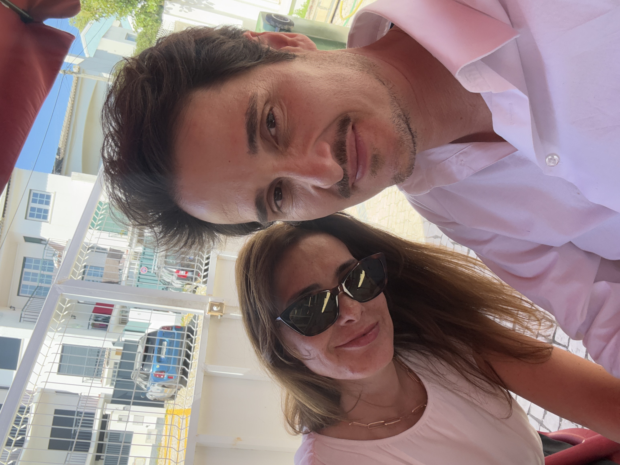 A selfie of a man and a woman in Cascais, Portugal. The man is on top, wearing a white button-down shirt. He has dark hair and a mustache. The woman is below him, wearing sunglasses and a white top with a gold chain necklace. A building with multiple windows and a blue car behind a white fence is visible in the background. The scene appears to be outdoors during daylight.