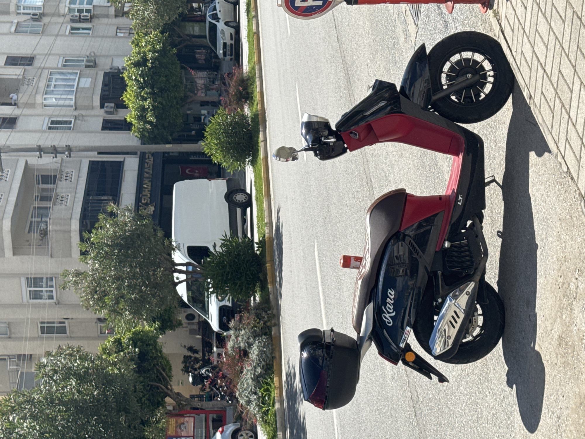 The image depicts a parked Kara scooter, predominantly black and red in color, situated on a paved area alongside a city street. The scooter has a rear storage compartment. Behind the scooter, there's a street with vehicular traffic, including a white van. Across the street is a building with multiple floors, trees, and some landscaping. The image suggests an urban environment during daylight hours.