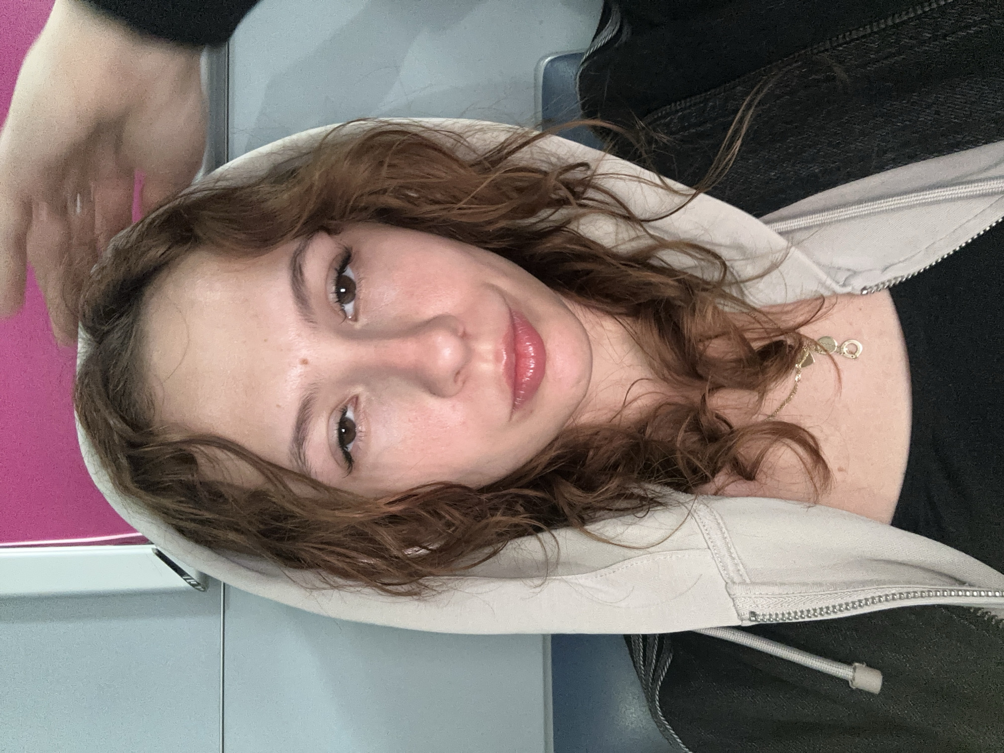 A woman with shoulder-length, curly brown hair, wearing a light beige hoodie and a black top, is positioned in what appears to be an interior space, possibly a bus or train, given the pink and grey wall behind her. Her face is in focus, with brown eyes and glossy lips. A glimpse of a gold necklace is visible around her neck. The image's orientation is sideways. There are no clear details suggesting the scene's location within Podgorica, Montenegro.