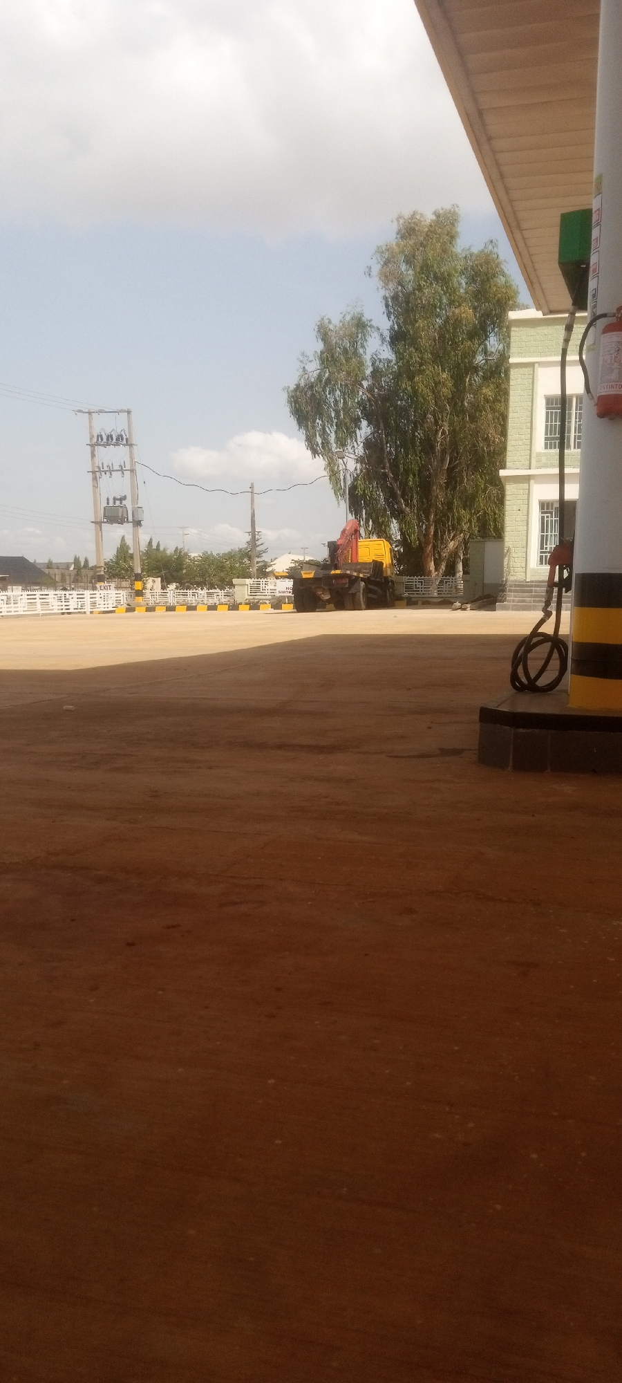 The scene depicts an open area, possibly a parking lot or a commercial space, in Sabon Tasha, Nigeria, on a sunny day. In the background, there's a yellow crane truck parked near a white fence with yellow and black striped posts. An electrical utility pole with transformers is visible further in the distance. A tree stands behind the crane. The foreground is predominantly a brown, dusty surface. To the right, a section of a fuel pump with black and yellow striping is visible, as is part of a building with green window frames and a fire extinguisher. The sky is partially cloudy.
