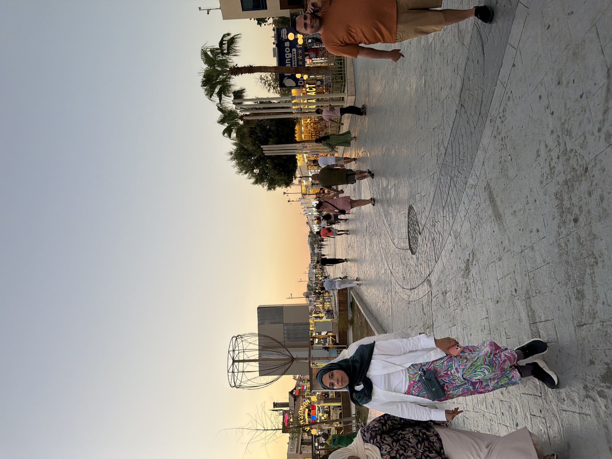 The image depicts a bustling pedestrian promenade in Sharm El Sheikh, Egypt, during the late afternoon or early evening, as indicated by the soft, warm light of the setting sun. The promenade is paved with textured gray cobblestones, with distinct circular patterns integrated into the design. Palm trees are scattered along the walkway, and there are shops and stalls lining one side, illuminated by warm, artificial lights.
In the foreground, a woman wearing a green hijab, a white jacket, and a vibrant, multi-colored patterned skirt is walking towards the viewer. She has a small, dark green crossbody bag and is holding a pink smartphone in her right hand. To her left, another woman in a patterned top and light-colored trousers is partially visible.
Further down the promenade, various individuals are walking in both directions. A man in an orange t-shirt and khaki shorts is on the right, speaking on his phone and looking towards the viewer with a smile. Other people are seen in groups and individually, some in casual summer attire. In the background, several structures and buildings are visible, including a distinctive metal sculpture resembling a large cage or basket, and storefronts with neon signs. The sky is a clear gradient of pale blue to orange, signifying the approaching sunset. The overall atmosphere appears lively and relaxed, characteristic of a popular tourist area.
